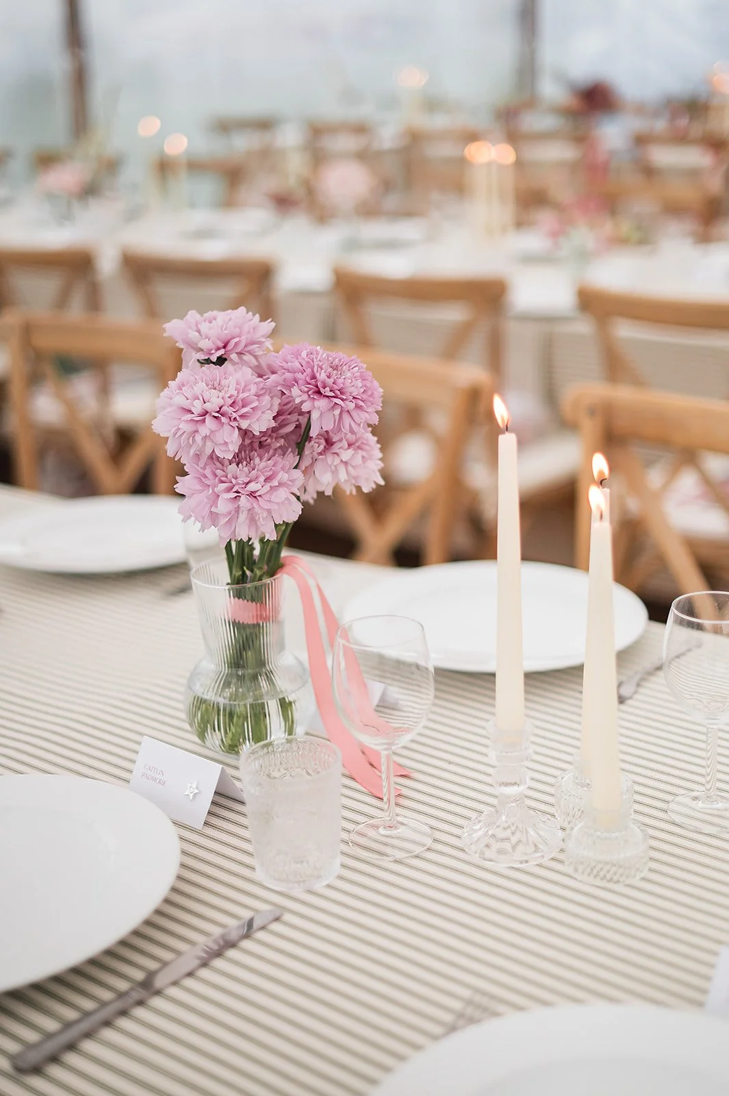 Elegant dining table set with pink flowers in a glass vase, lit candles, white plates, wine glasses, and silverware, in a well-lit room with wooden chairs.