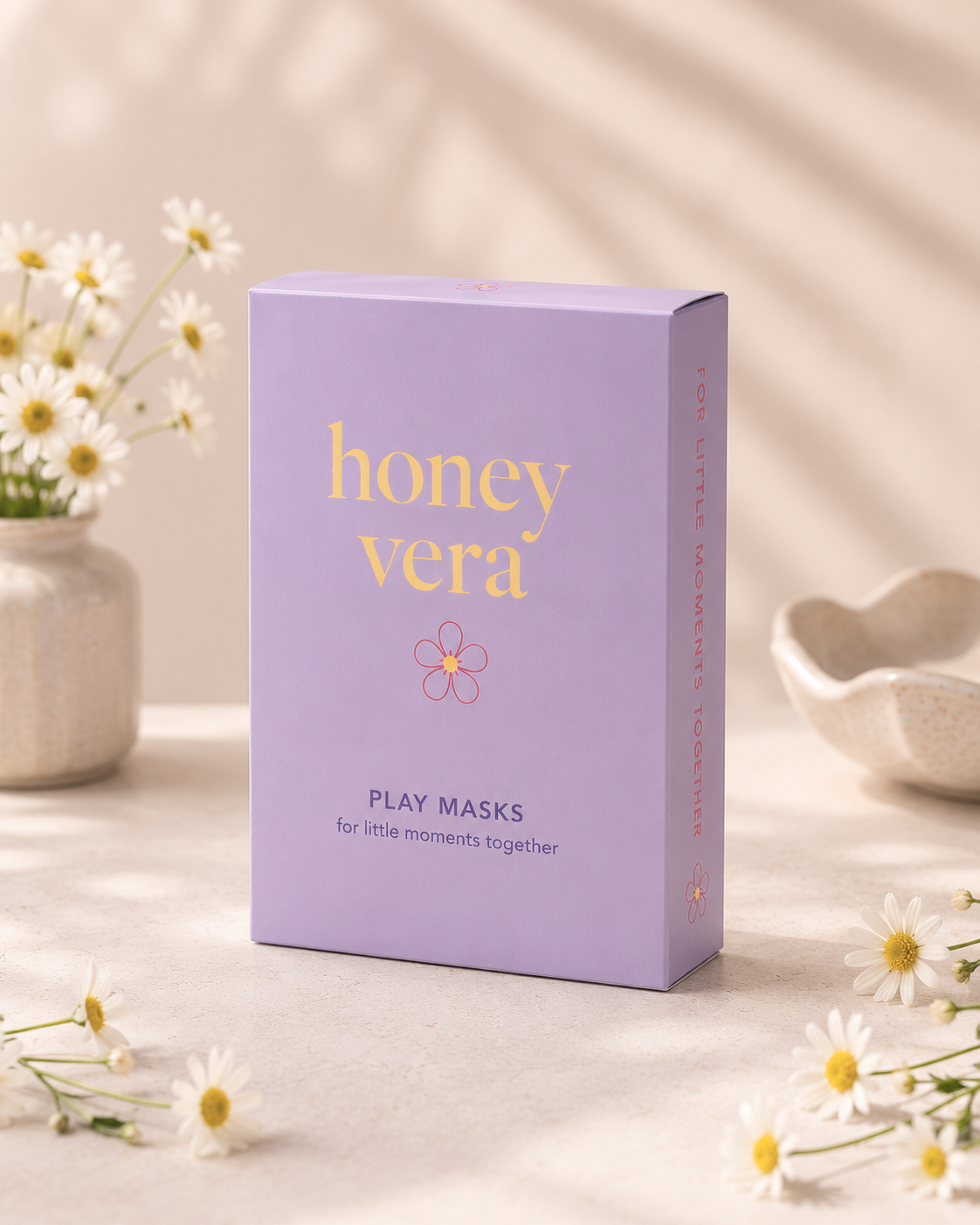 A lavender box labeled 'honey vera' with yellow and purple text, and a small flower illustration, is on a light surface with small daisies and ceramic bowls around it.