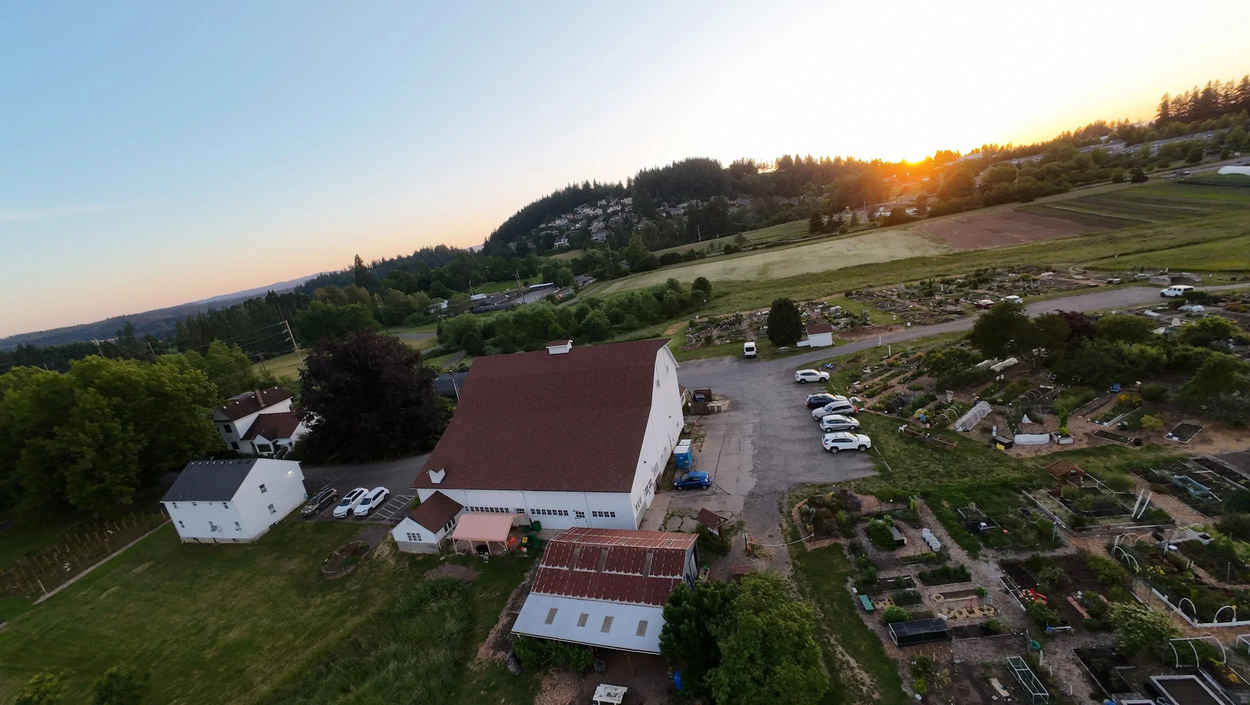 Aerial view of a rural area at sunset with houses, cars, green fields, and trees.