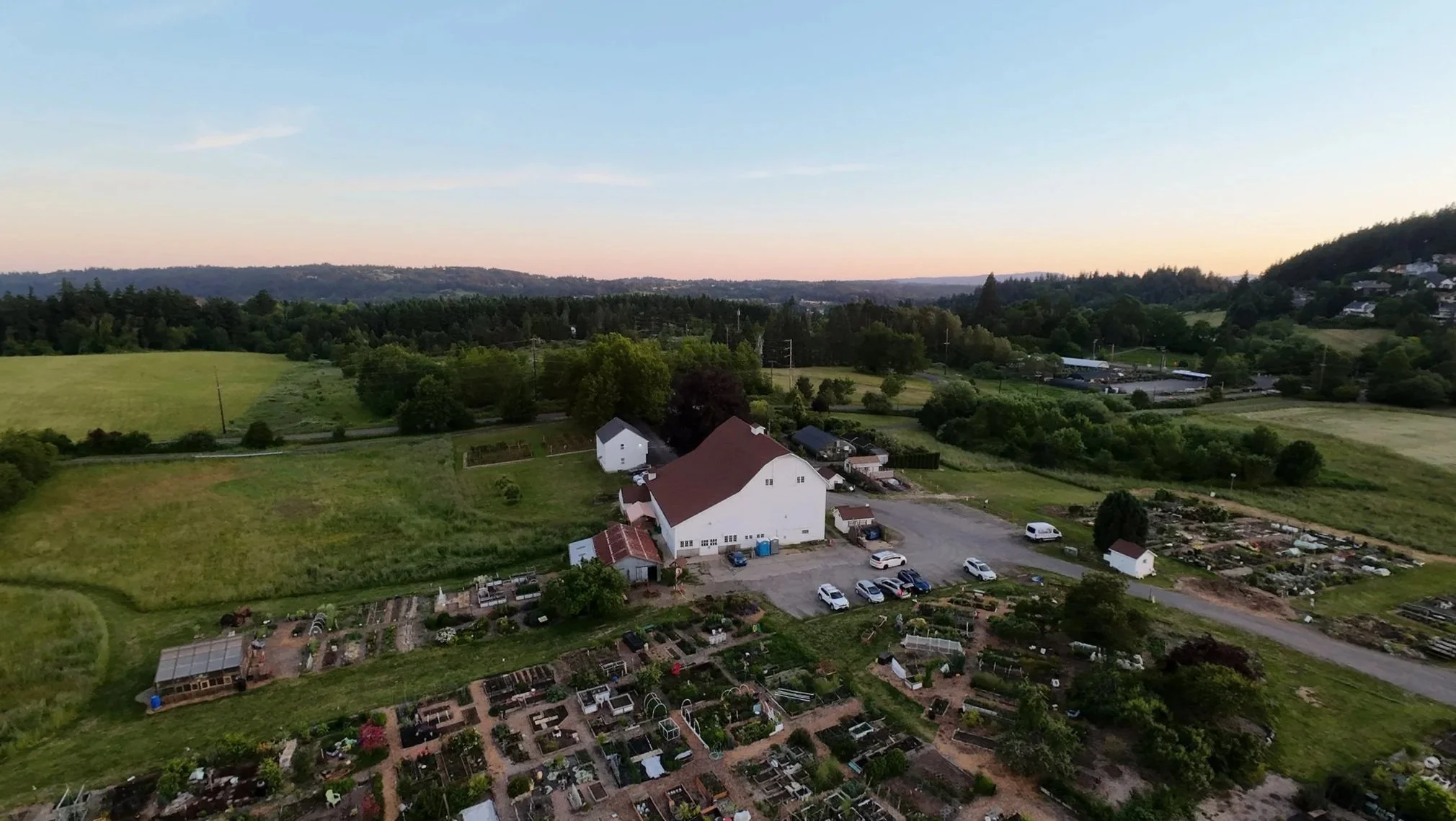 A rural landscape during sunset with a large white barn, surrounding green fields, and multiple houses on a hillside, under a clear sky.