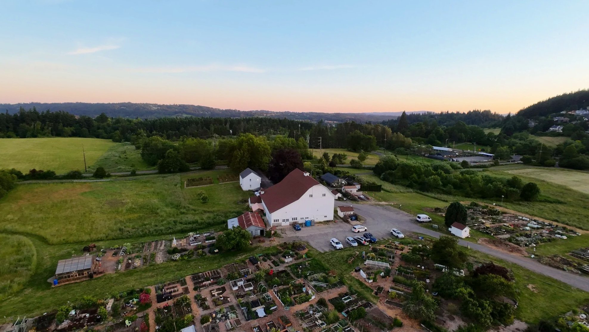 Aerial view of a rural landscape Luscher Farm in Lake Oswego with green fields, trees, houses, and a large white barn, during sunset with a clear sky.