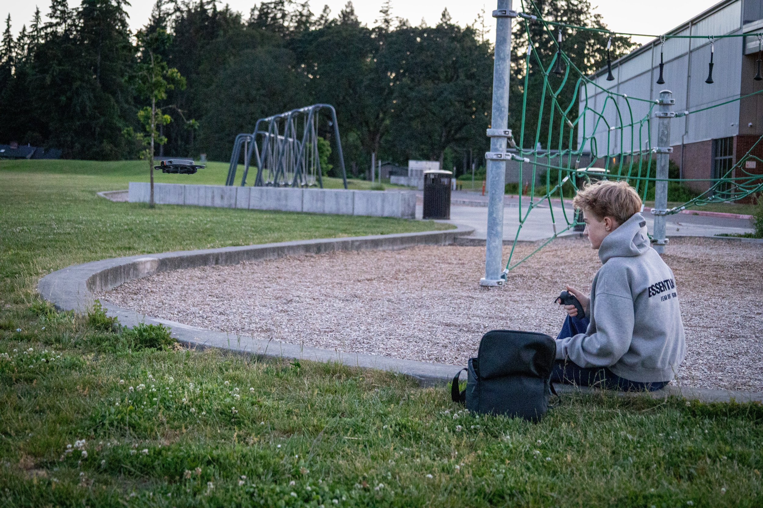 Henry Lavoie sitting on the ground holding a remote control, looking at a drone flying near a playground in a park during dusk.