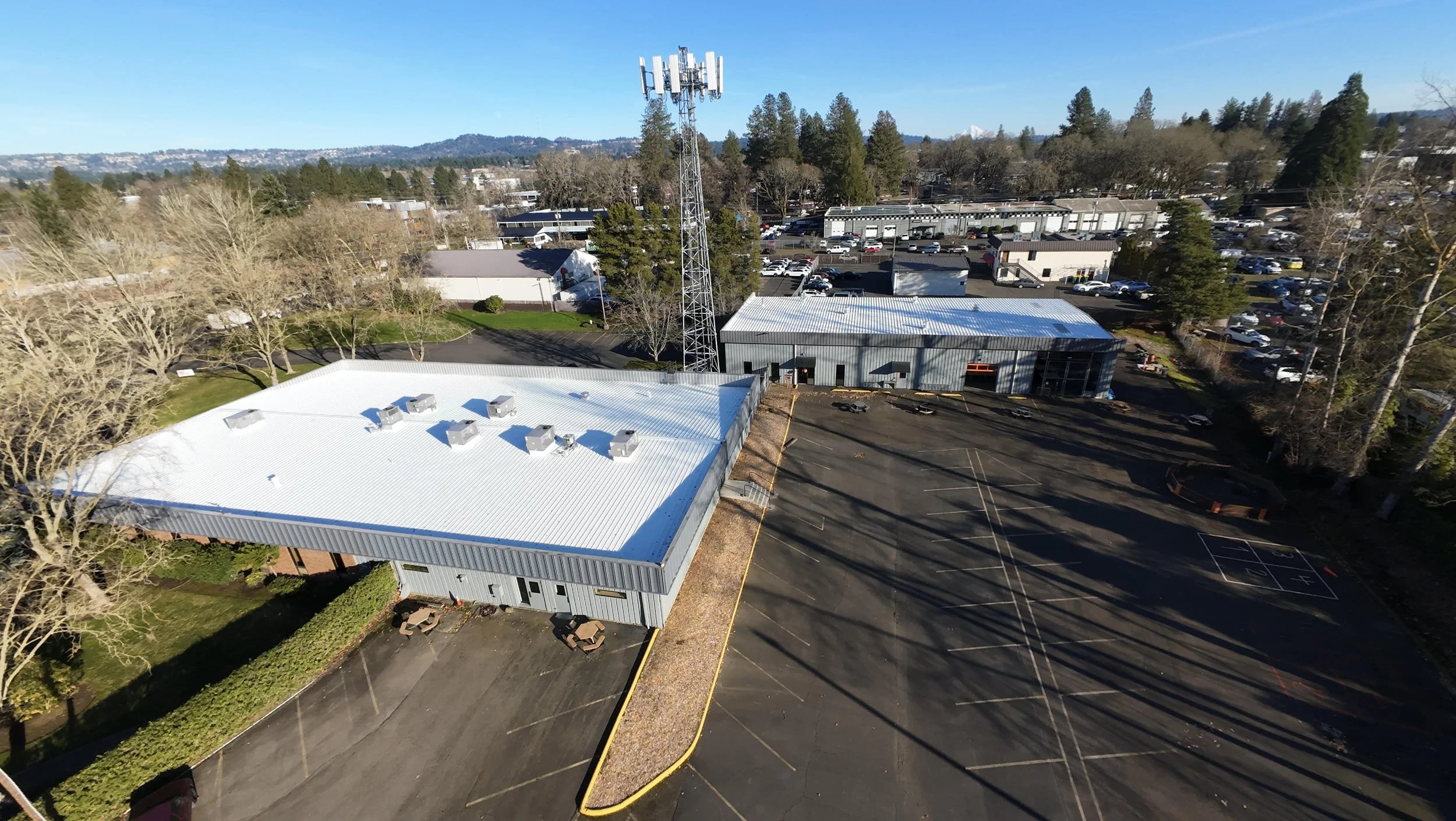 Aerial view of a parking lot, several buildings, and a cell tower surrounded by trees under a clear blue sky.