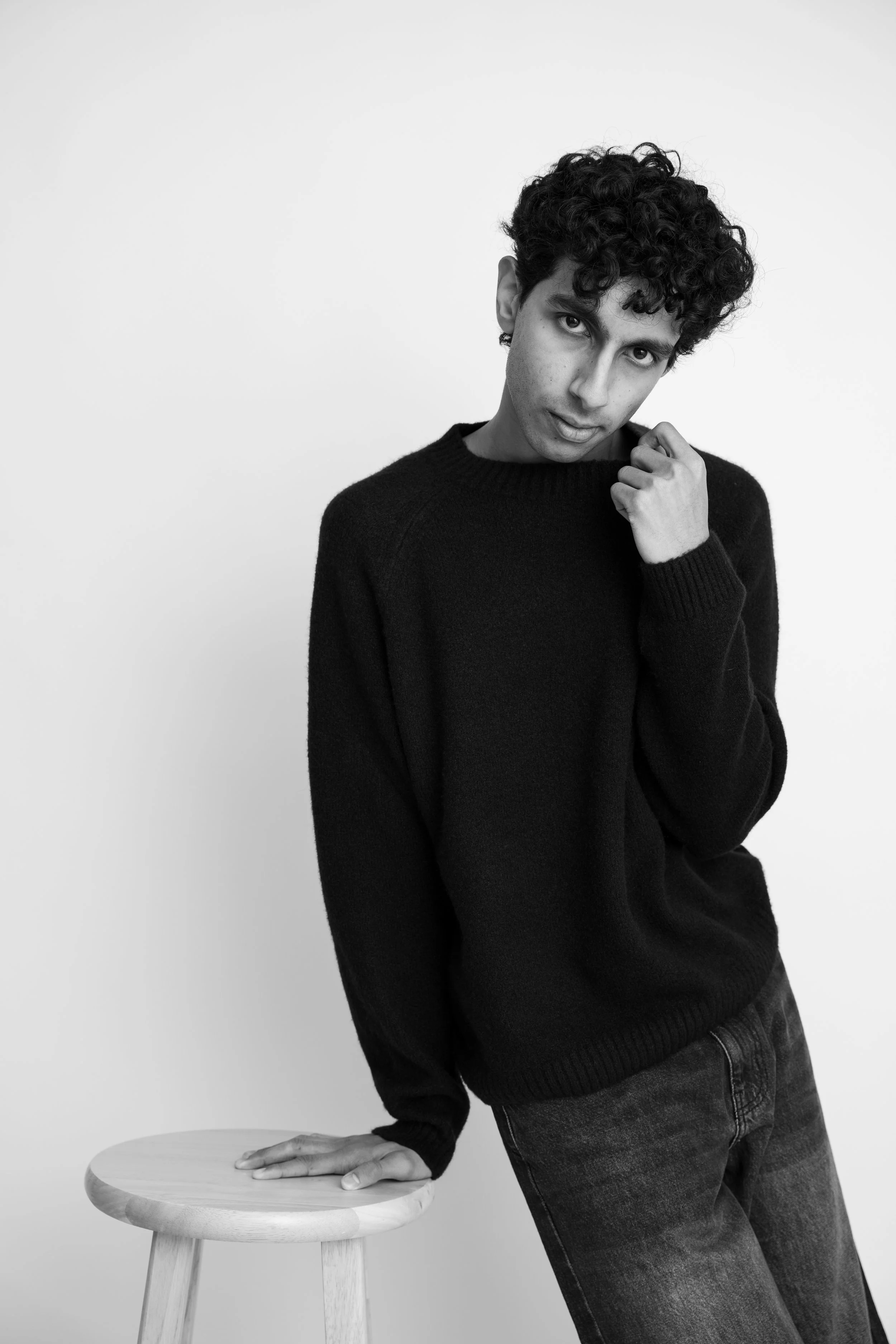A young man with curly hair and light skin, wearing a black sweater and dark jeans, leaning on a small wooden stool with one hand, in front of a plain white background, looking at the camera with a slightly tilted head.