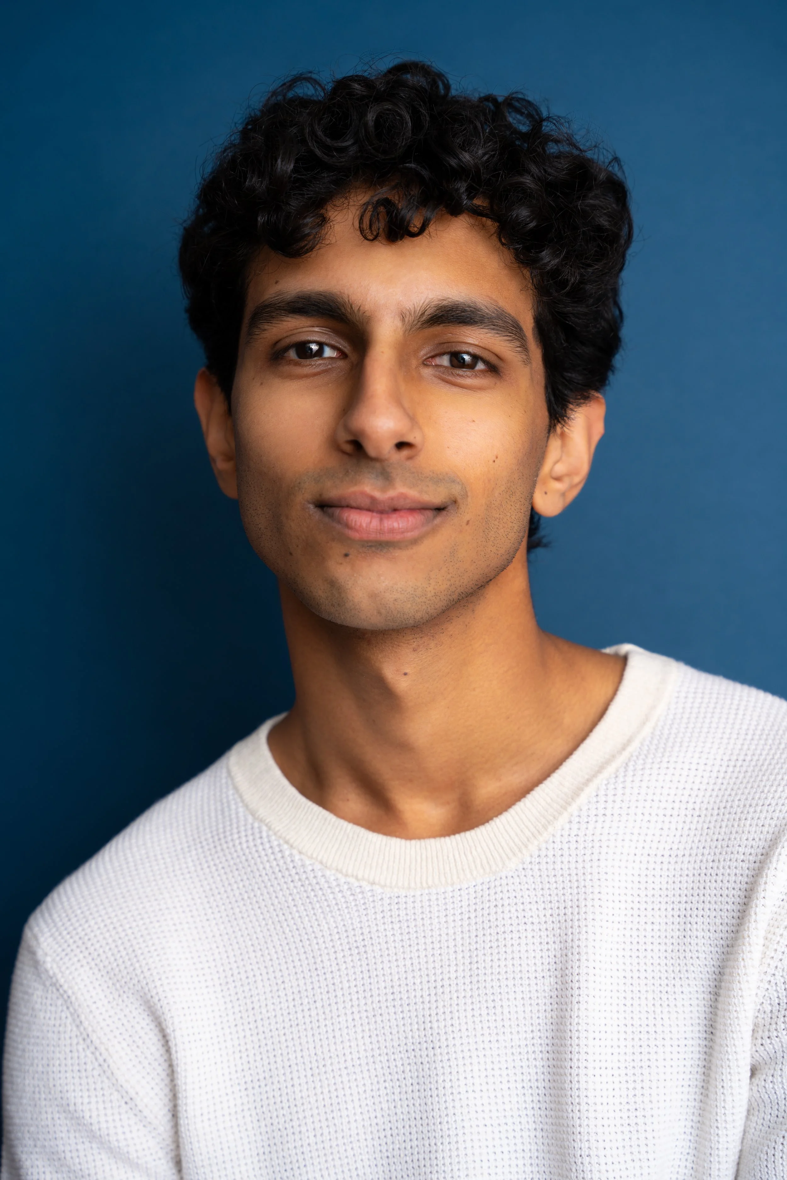 Close-up of a young man's face with short curly black hair, wearing a white textured sweater, against a solid dark blue background.