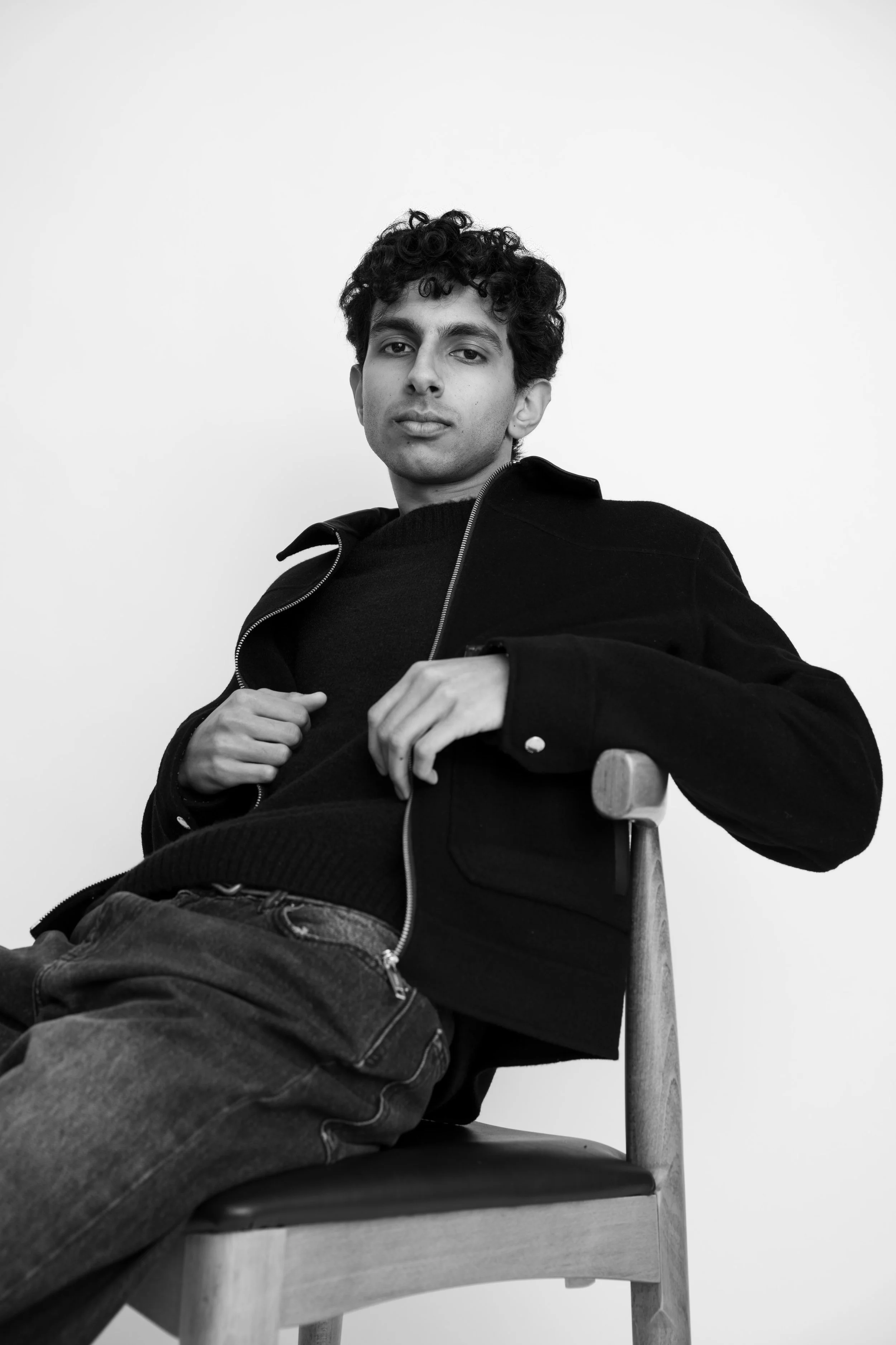 Black and white photo of a young man with dark curly hair sitting on a wooden chair with a relaxed posture, wearing a dark jacket and jeans, looking directly at the camera against a plain light background.