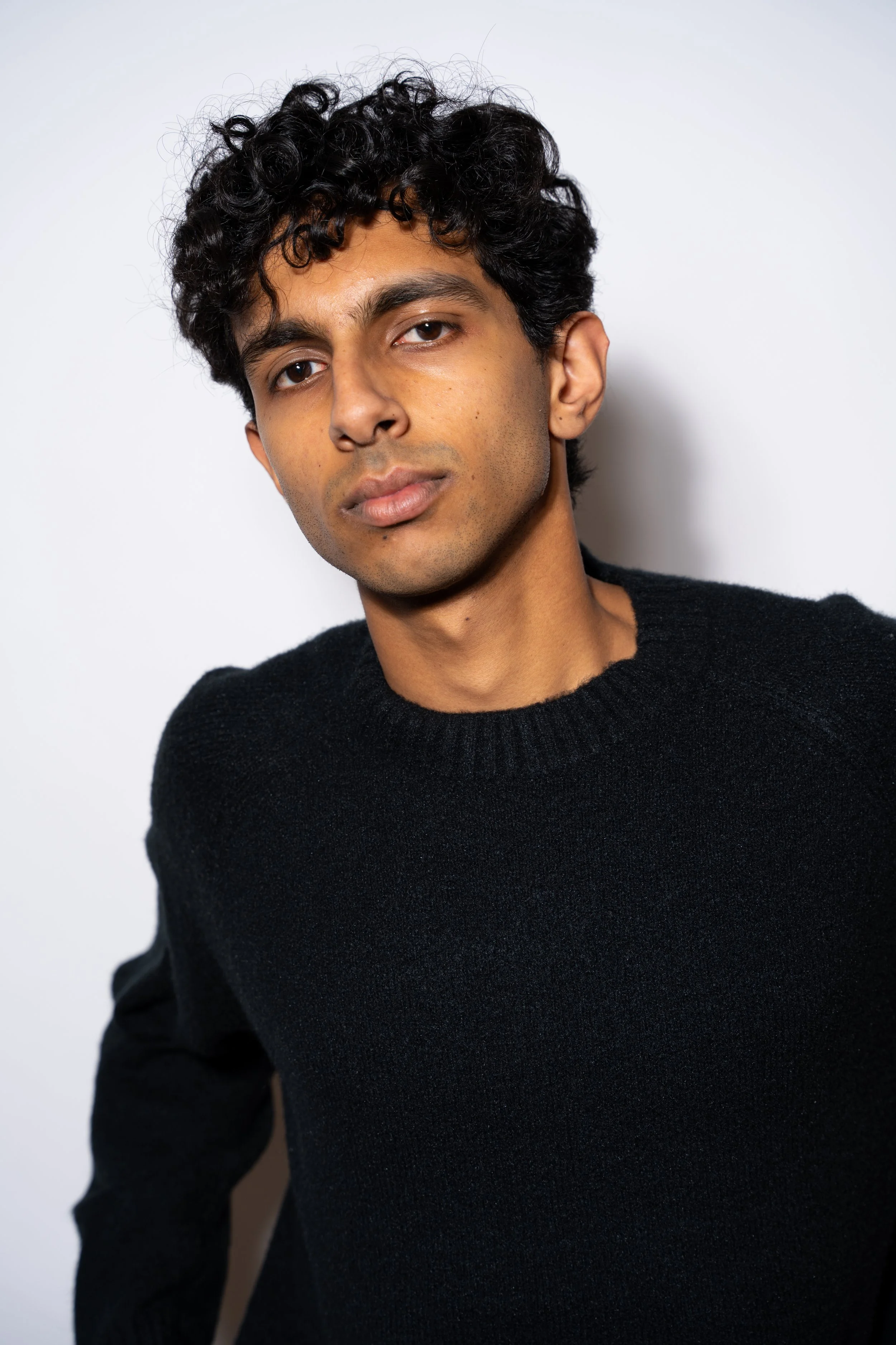 A young man with dark, curly hair and medium skin tone wearing a black sweater against a plain white background.