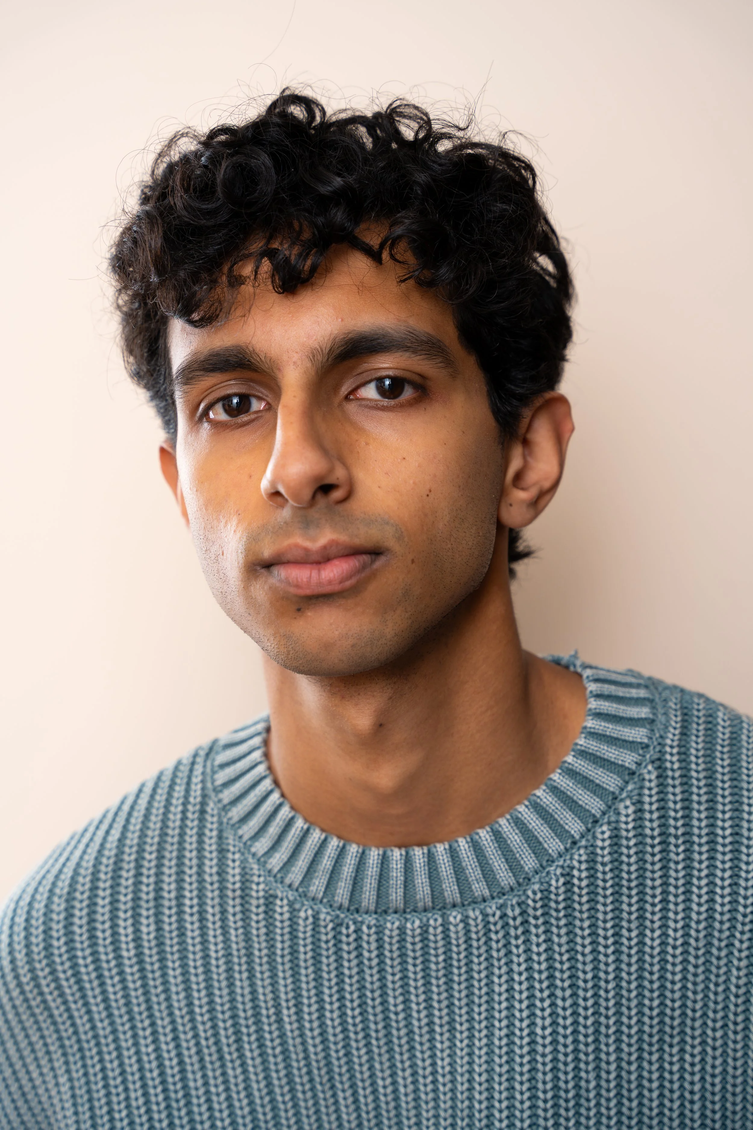 Close-up of a young man with short, curly dark hair, wearing a striped blue and gray sweater, against a neutral background.