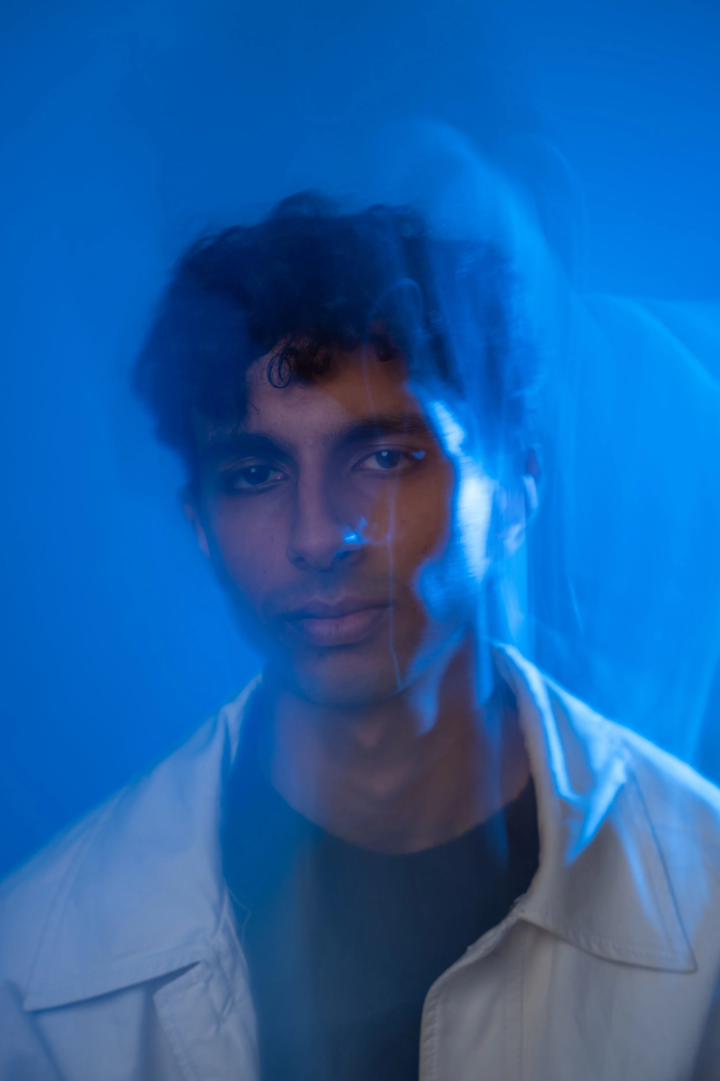 A young man with dark curly hair looking directly into the camera, wearing a white shirt, with a blue background and blue lighting creating a soft, mysterious effect.
