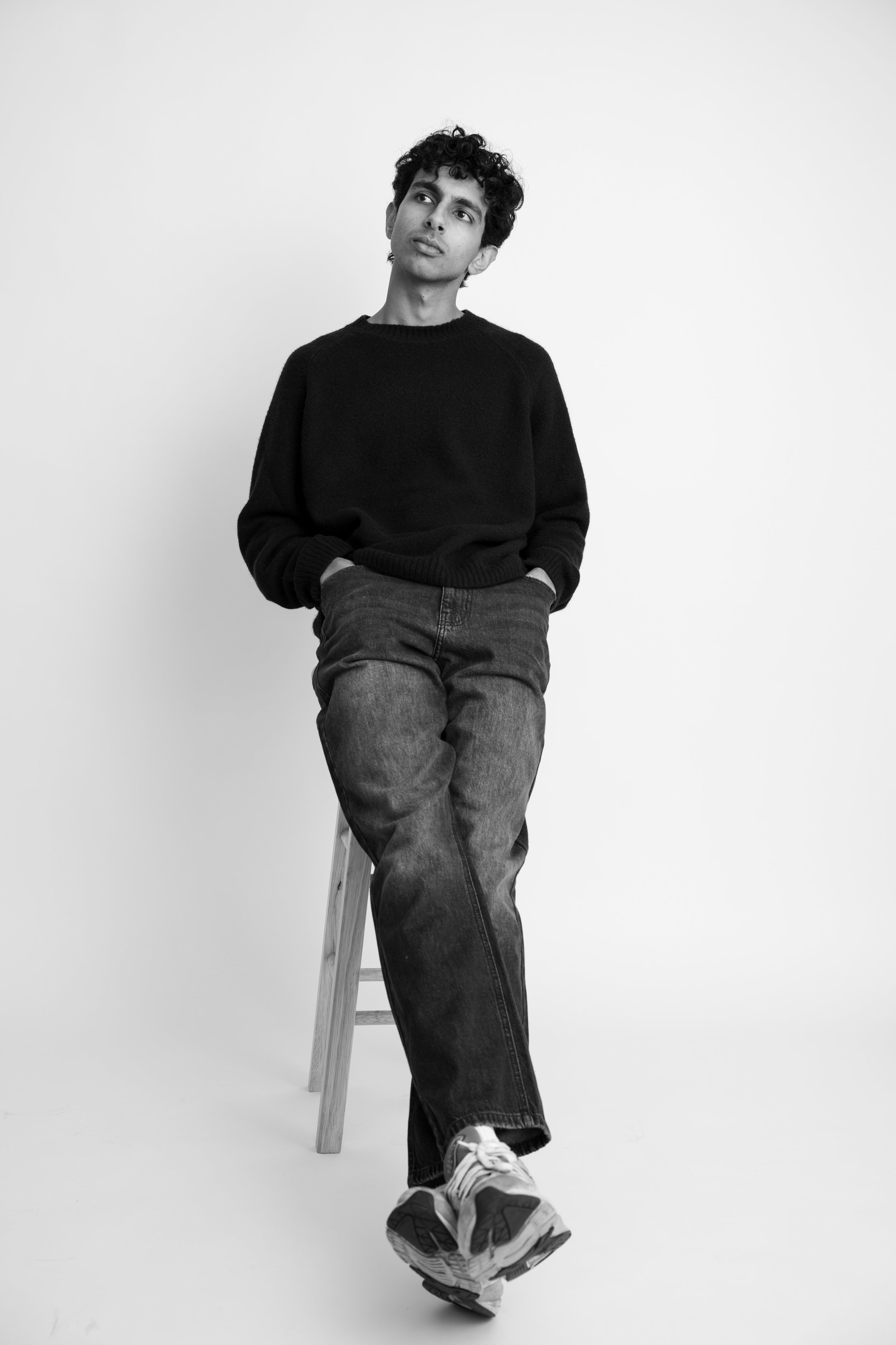 A young man with curly hair, dressed in a black sweater and jeans, seated on a wooden stool with one foot resting on the floor and the other slightly elevated, against a plain white background.