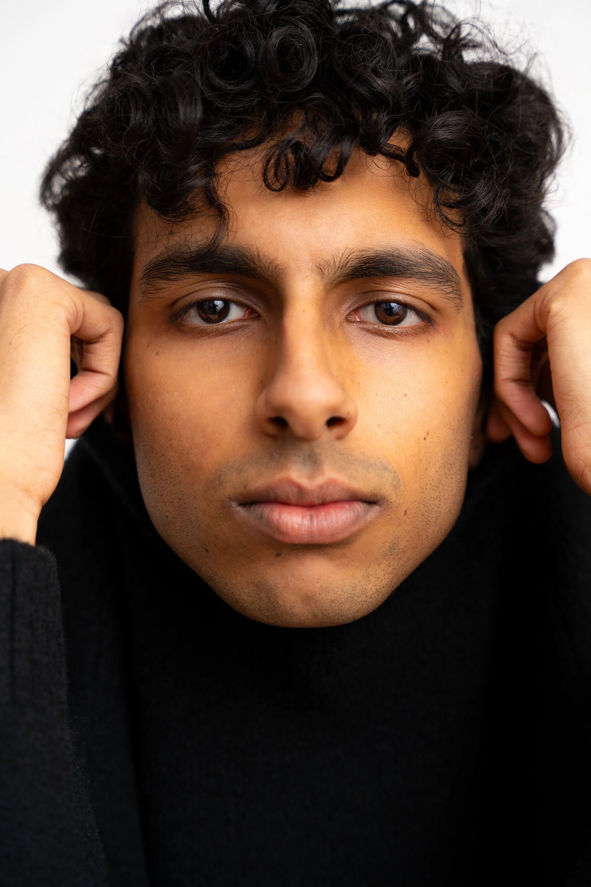 Close-up of a young person with curly dark hair, brown eyes, and light brown skin, wearing a black sweater, holding their hands near their ears, looking directly at the camera.