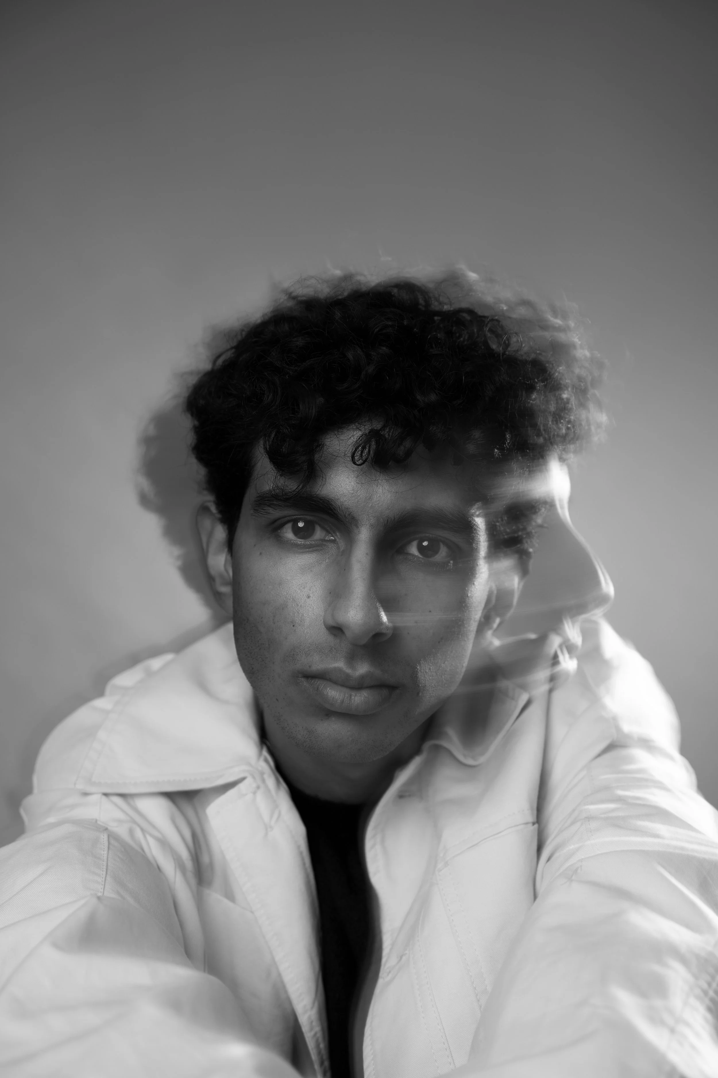 Black and white photo of a young man with curly hair, wearing a white shirt, looking directly at the camera with a neutral expression.