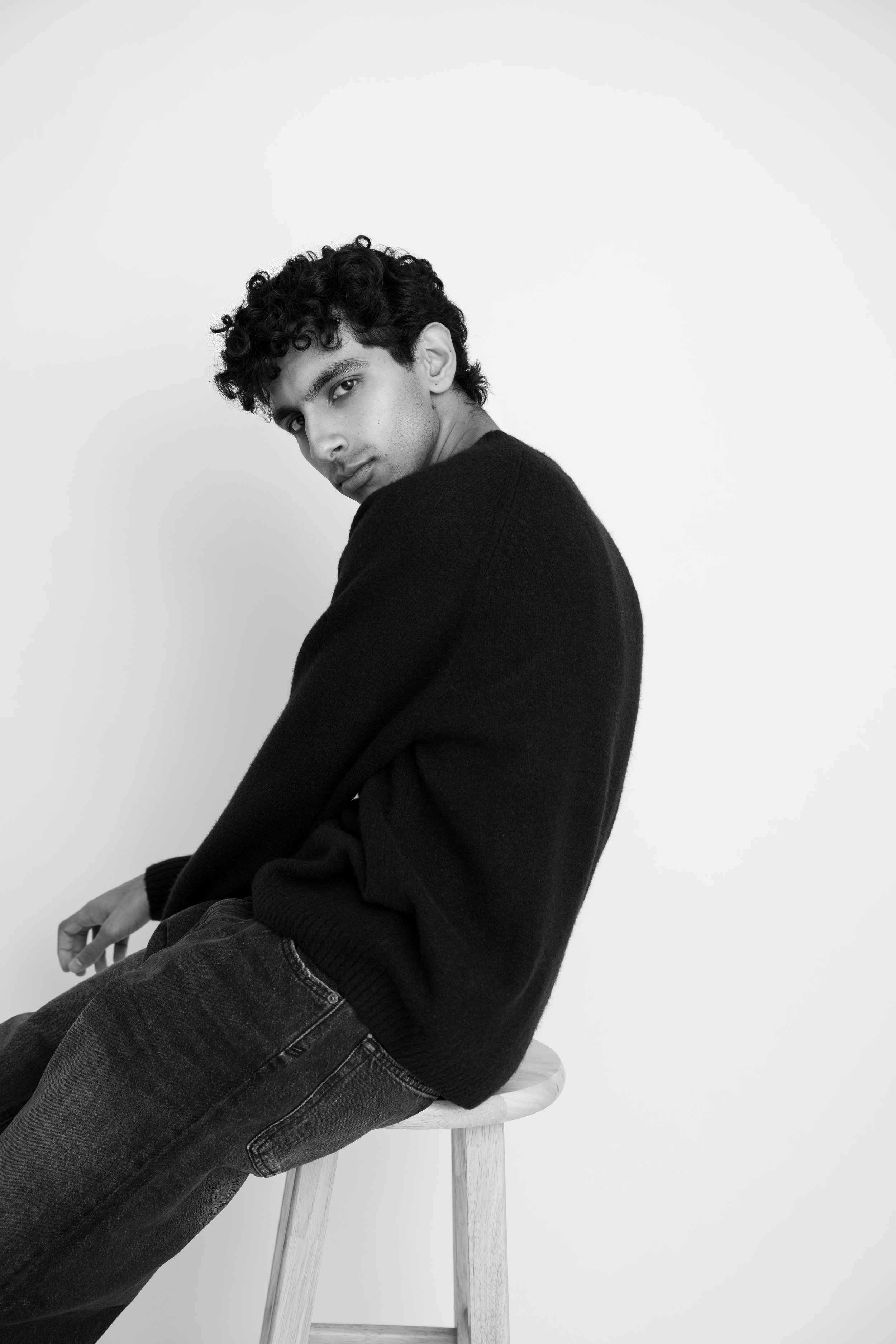 Black and white photo of a young man with curly hair sitting on a stool, wearing a black sweater and jeans, looking over his shoulder at the camera against a plain white background.