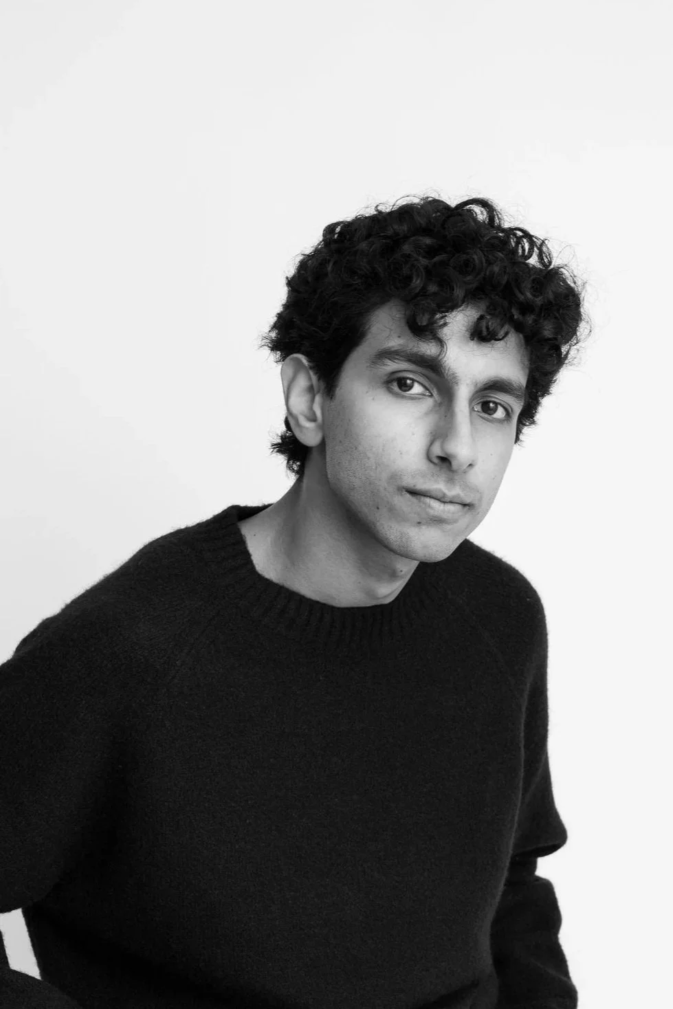 Black and white portrait of a young man with curly hair wearing a dark sweater, looking at the camera with a neutral expression, against a plain white background.