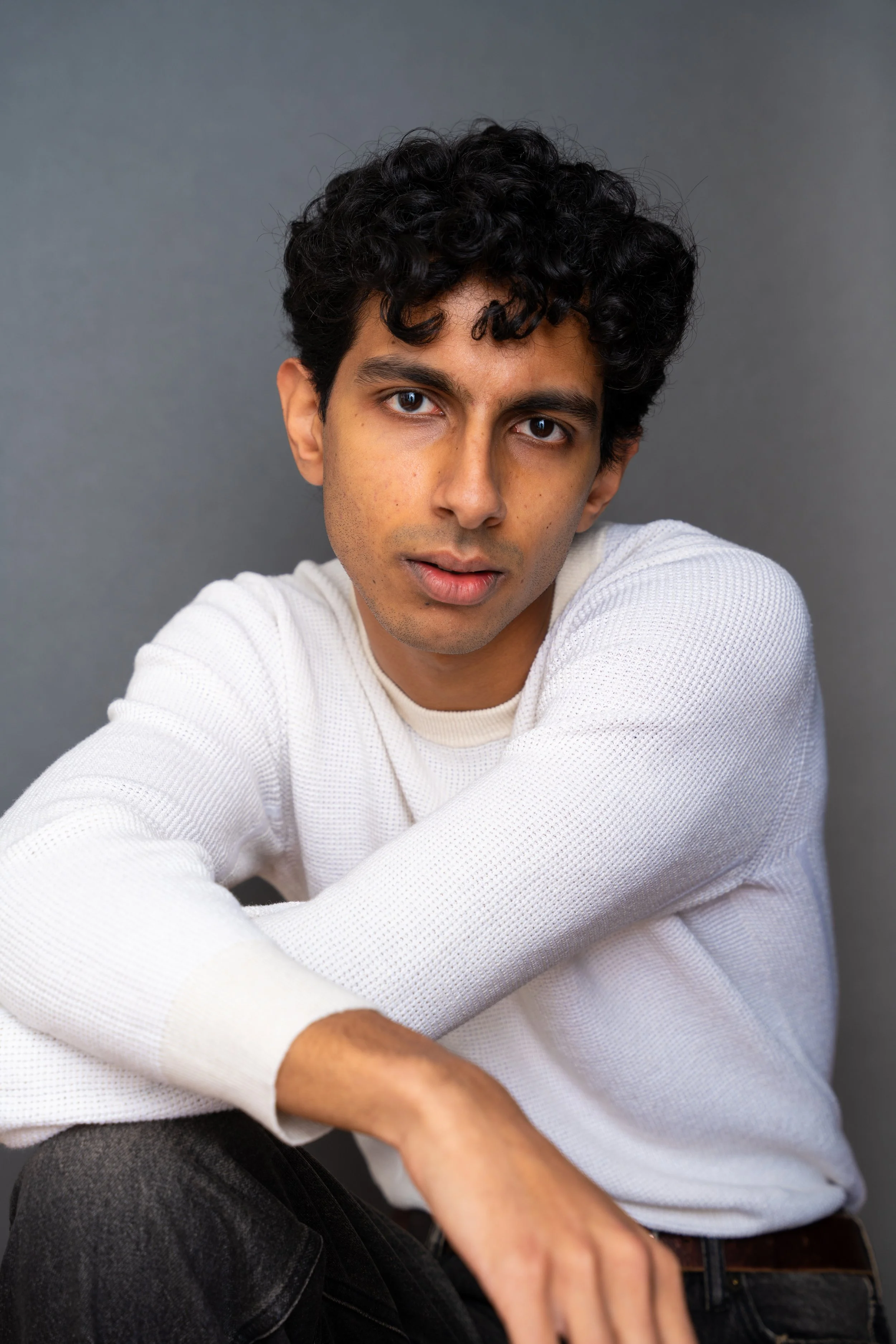A young man with dark curly hair and brown eyes, wearing a white textured long-sleeve shirt and black jeans, sitting against a plain gray background.