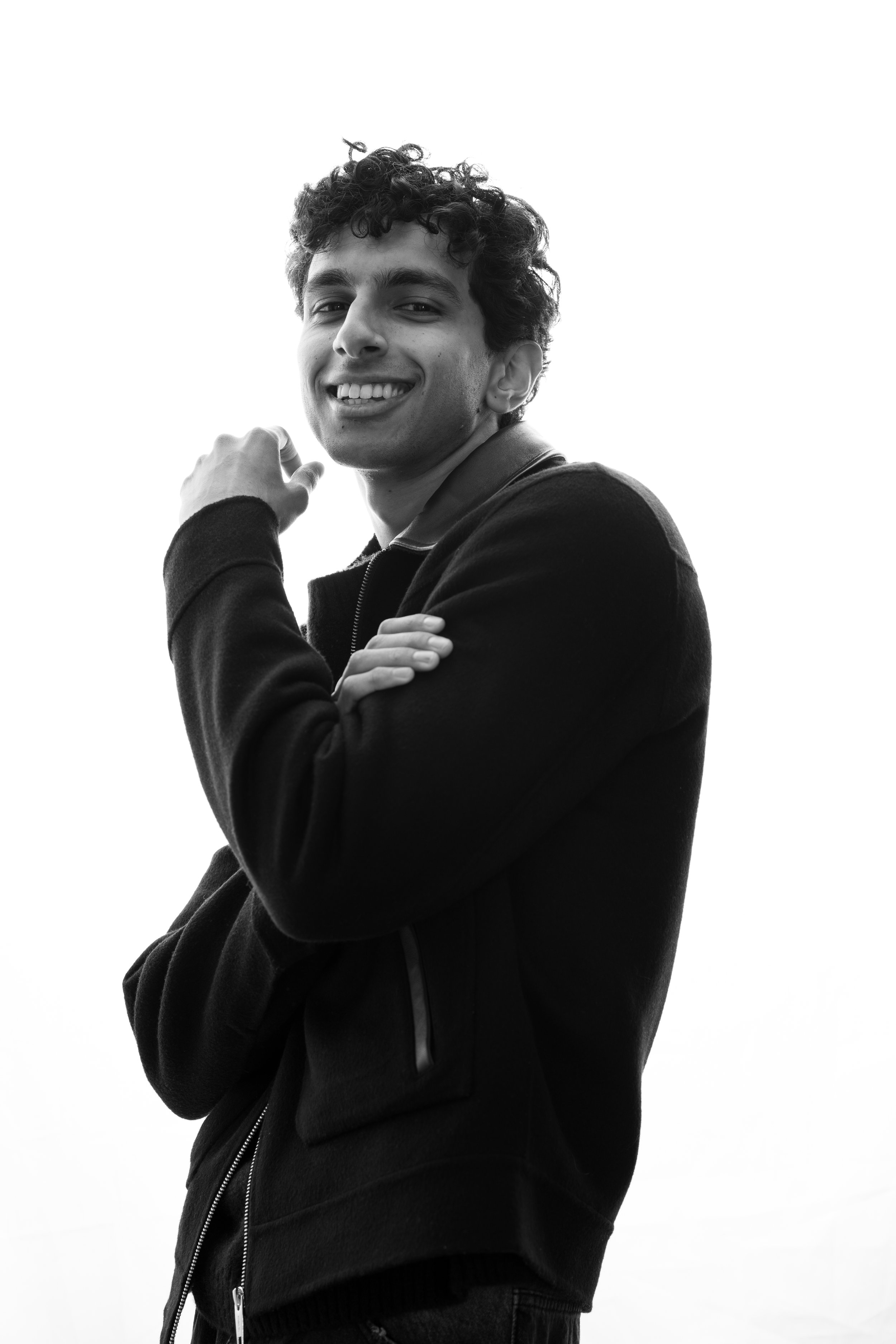 A young man with curly hair smiling and posing with his arm crossed, wearing a zip-up jacket, in black and white.