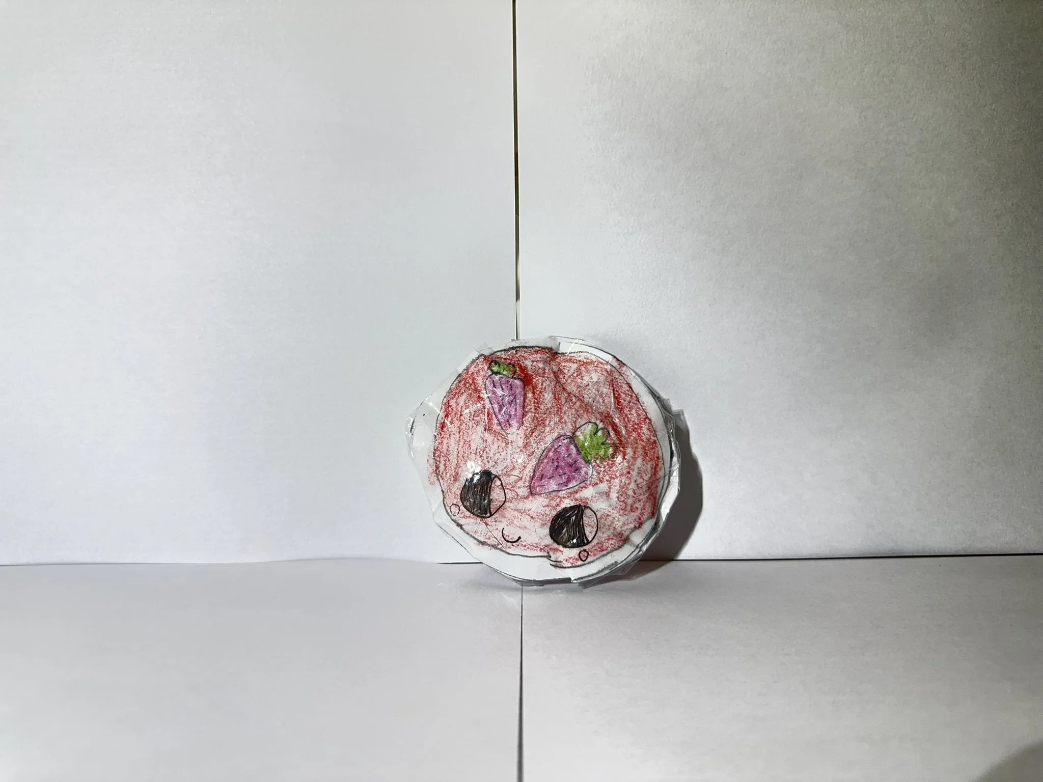 Children's drawing of a smiling strawberry with pink coloring and purple details, placed on a white surface.