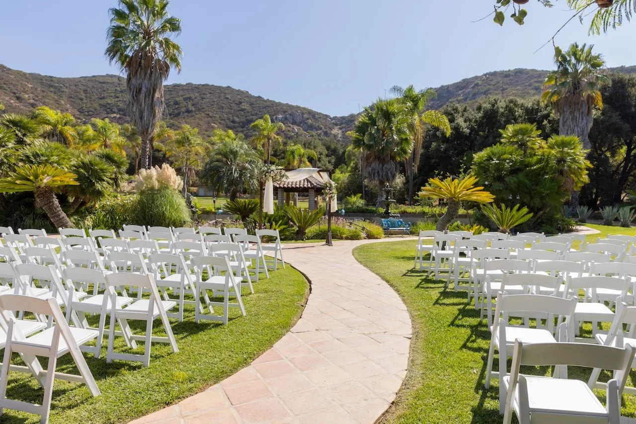 An outdoor wedding setup with white chairs arranged in rows along a curved pathway, surrounded by lush tropical plants and tall palm trees, with mountains in the background on a sunny day.