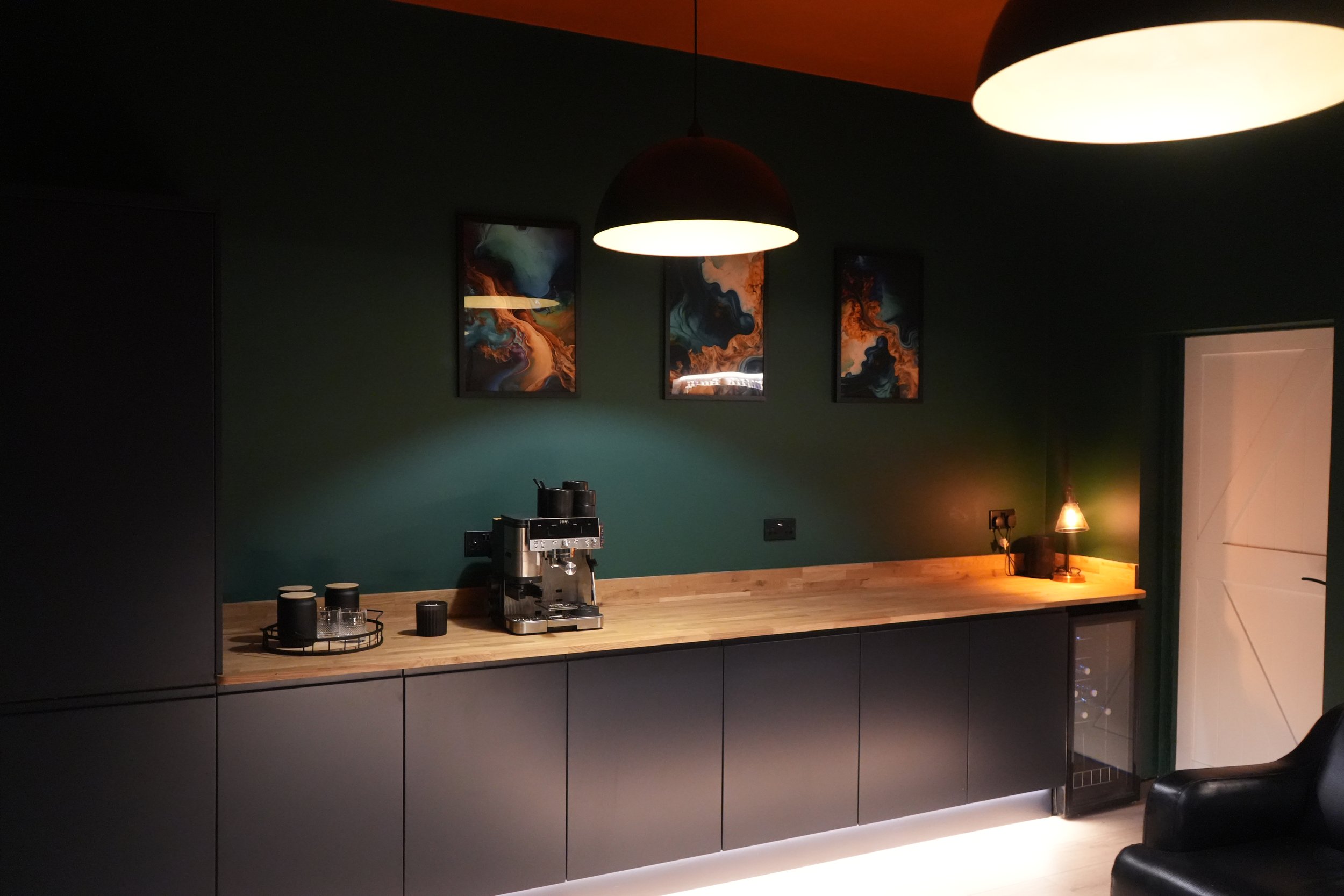 A dark countertop with a coffee machine, black cups, and glasses on a tray. Above, three colorful abstract art pieces hang on a green wall. Two large pendant lights hang from the ceiling, casting light on the counter. A small table lamp with a brass base and a glass shade is on the right. The overall atmosphere is cozy and modern.
