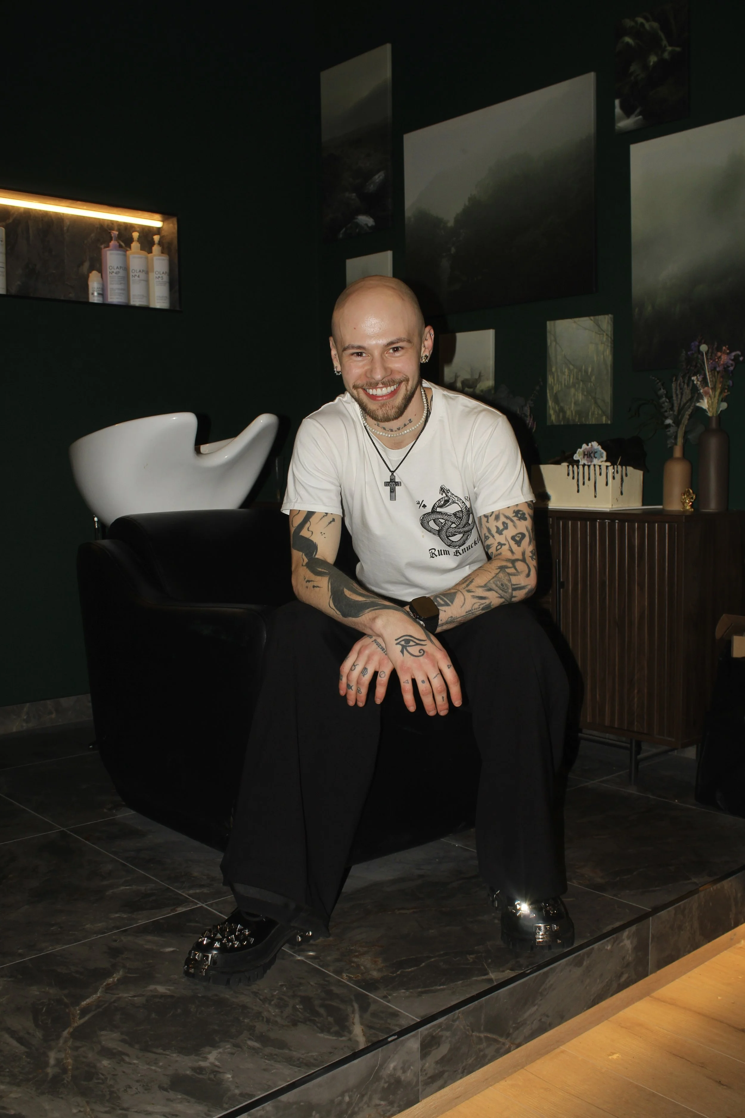 A smiling, tattooed man with a beard and piercings, wearing a white graphic t-shirt with a snake design, sitting in a modern lounge with dark walls and artwork, with a vase of flowers and a cake in the background.