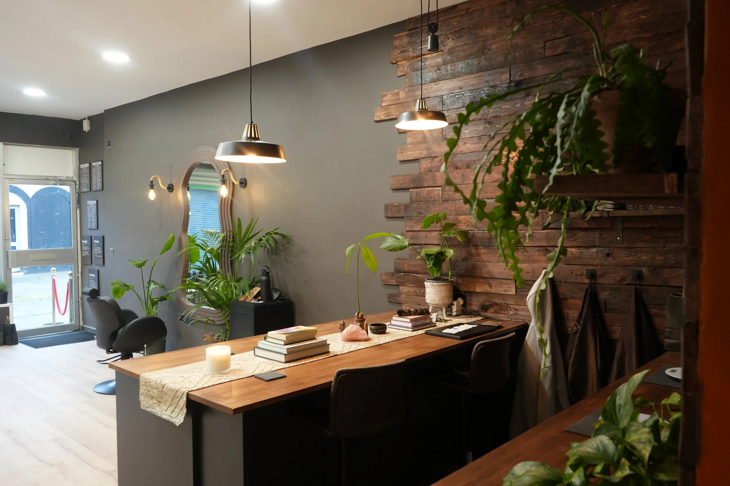 Interior of a modern salon with a wooden accent wall, plants on the table, hanging pendant lights, and chairs near the entrance.