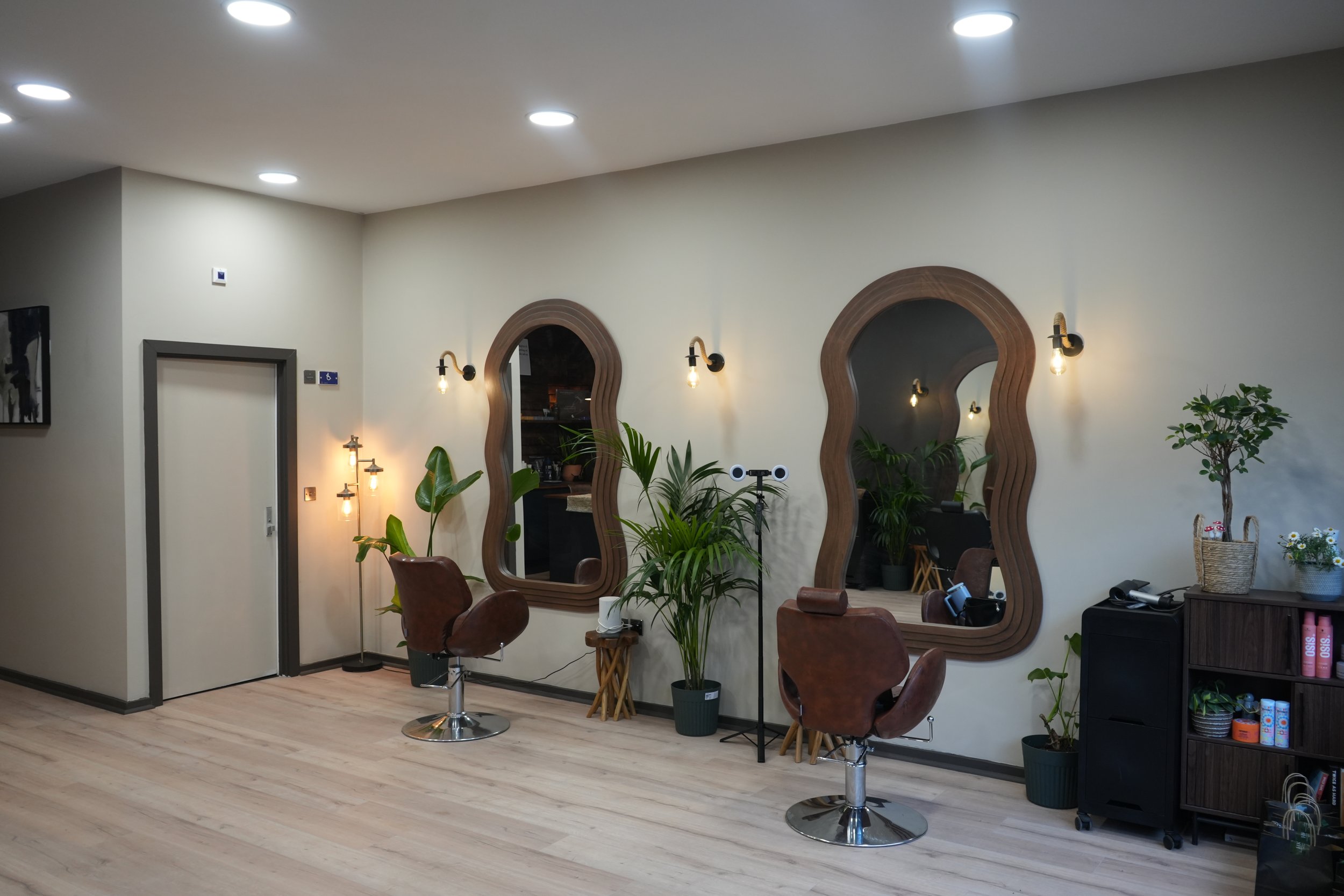 Interior view of a hair salon with two styling chairs, wooden mirrors, potted plants, and modern lighting.