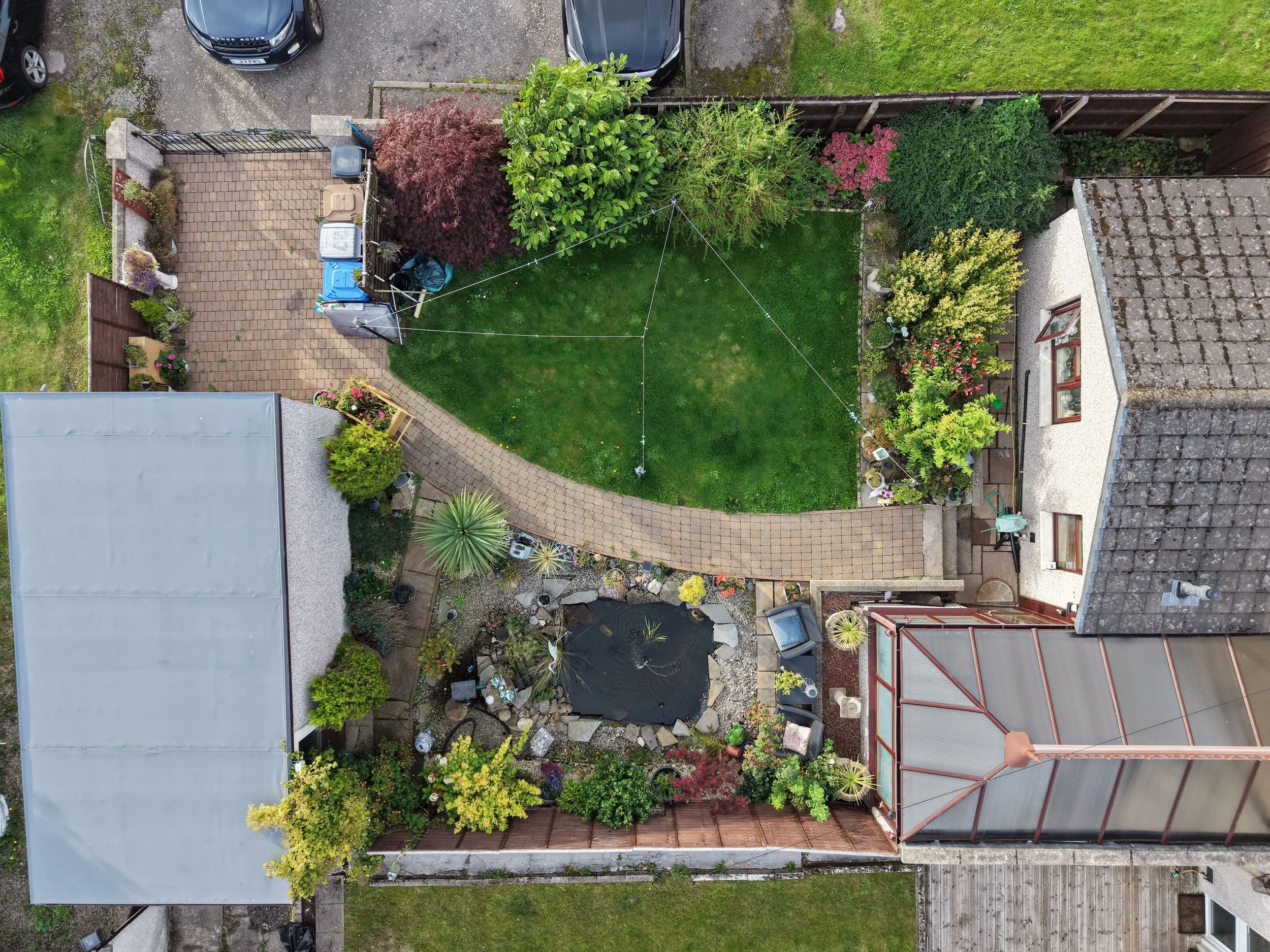 An aerial view of a backyard garden area with a lawn, various plants and trees, a paved pathway, a small pond, and outdoor decorations, bordered by neighboring buildings and parked cars.