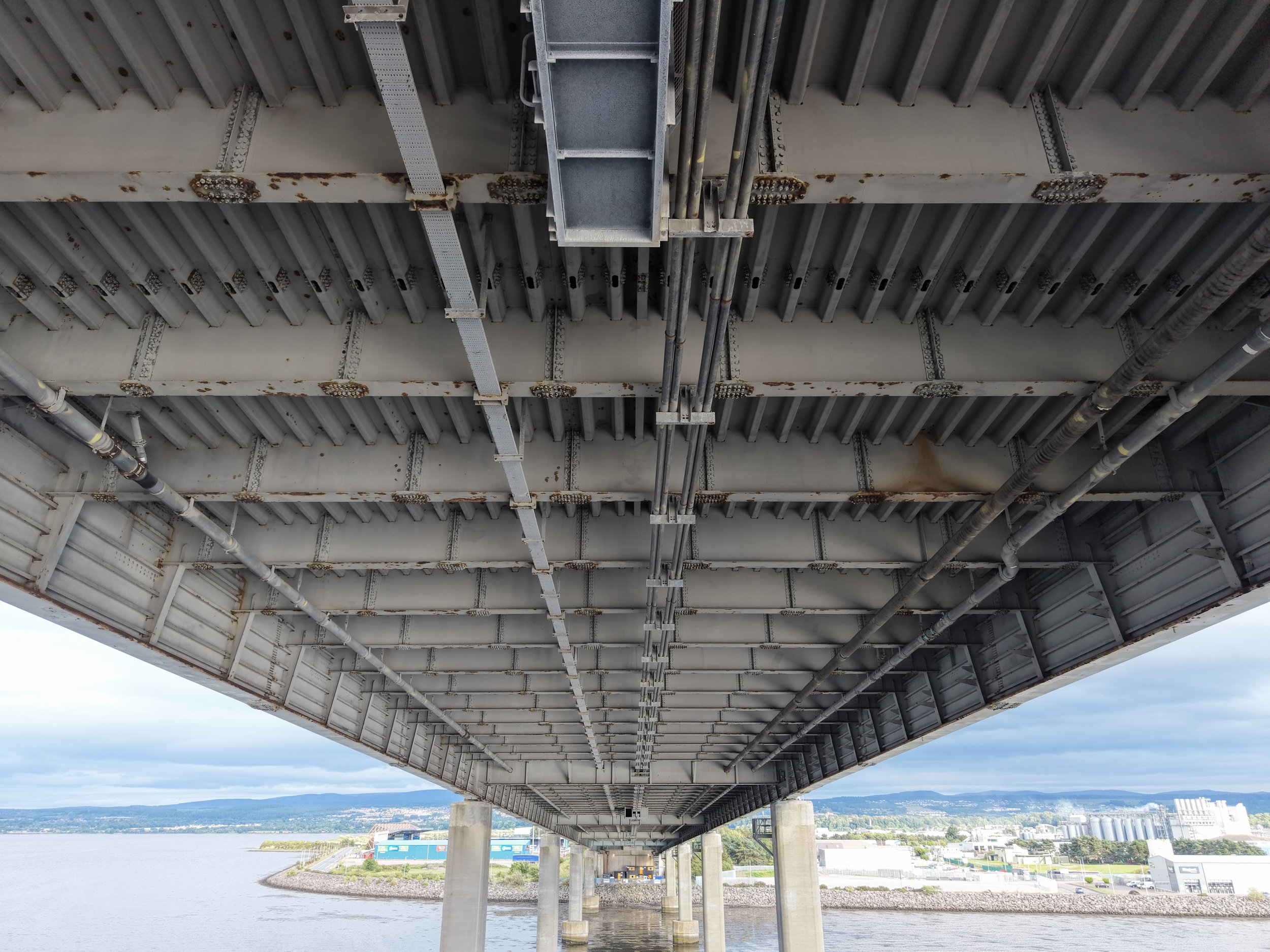 An underside view of a large bridge over a body of water, showing metal beams, pipes, and structural supports with a distant landscape of buildings and mountains.