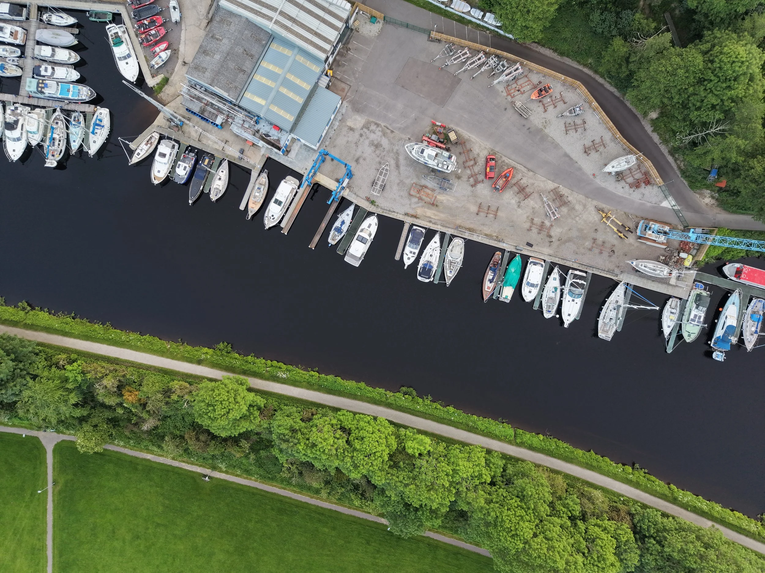 An aerial view of a marina with numerous boats docked along the water, adjacent to a construction area with boats on land, construction materials, and equipment, bordered by green trees and grassy areas.