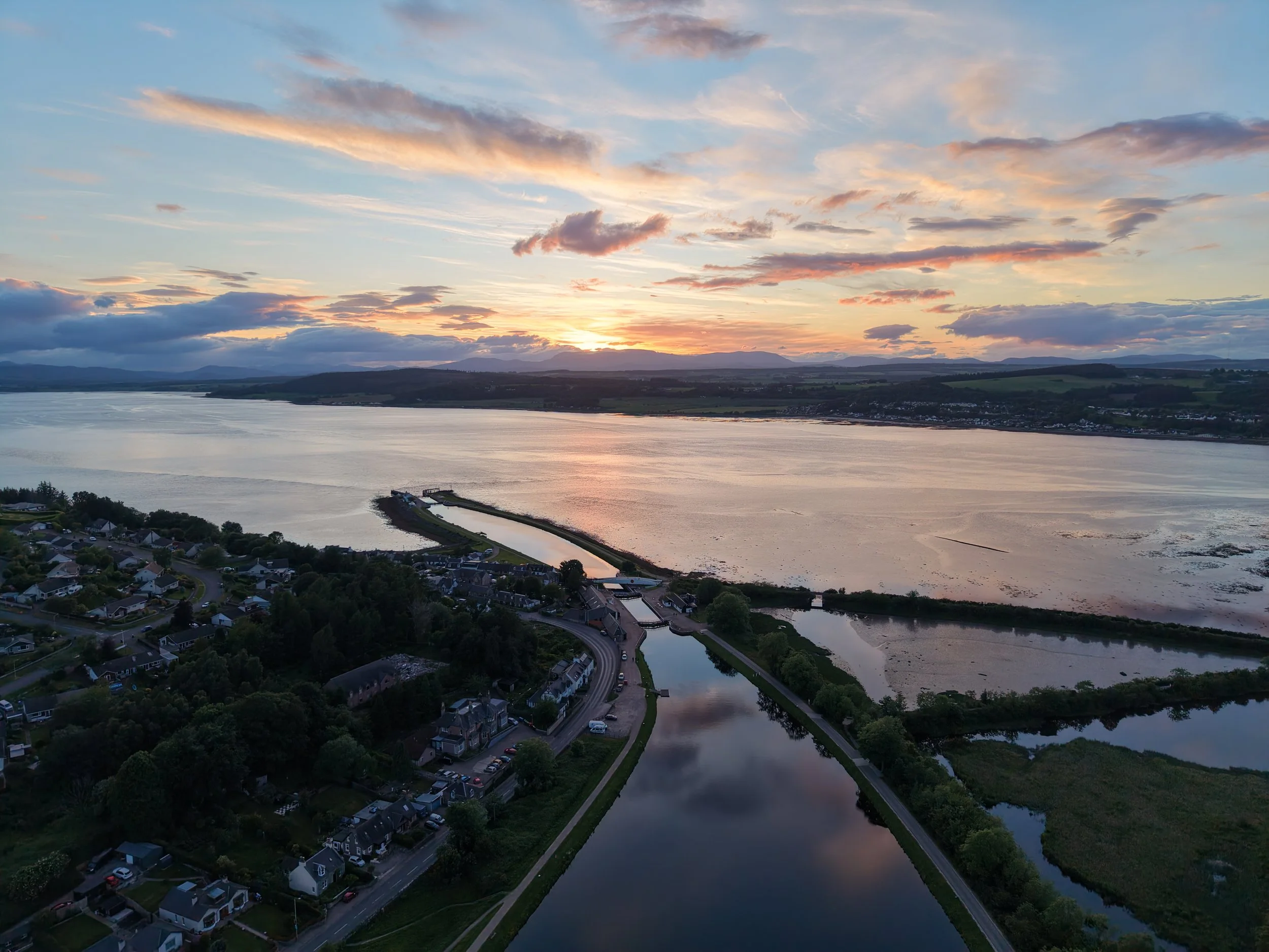 An aerial view of a landscape during sunset showing a river, residential houses, and a sky with scattered clouds.