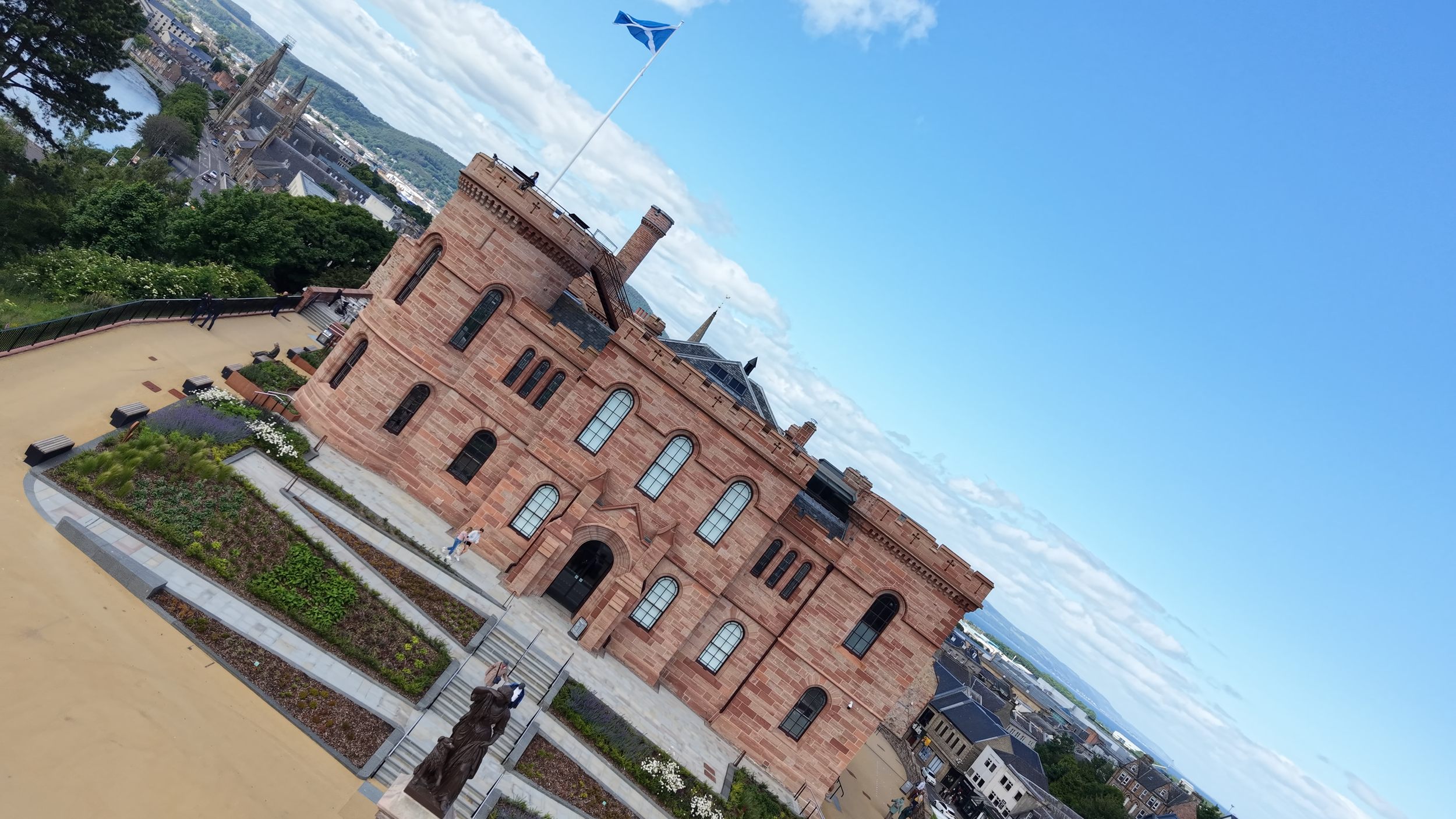 Historical red sandstone building with gothic windows, turrets, a flag on the roof, and a sculpture in front, set on a hill with cityscape and river in the background.