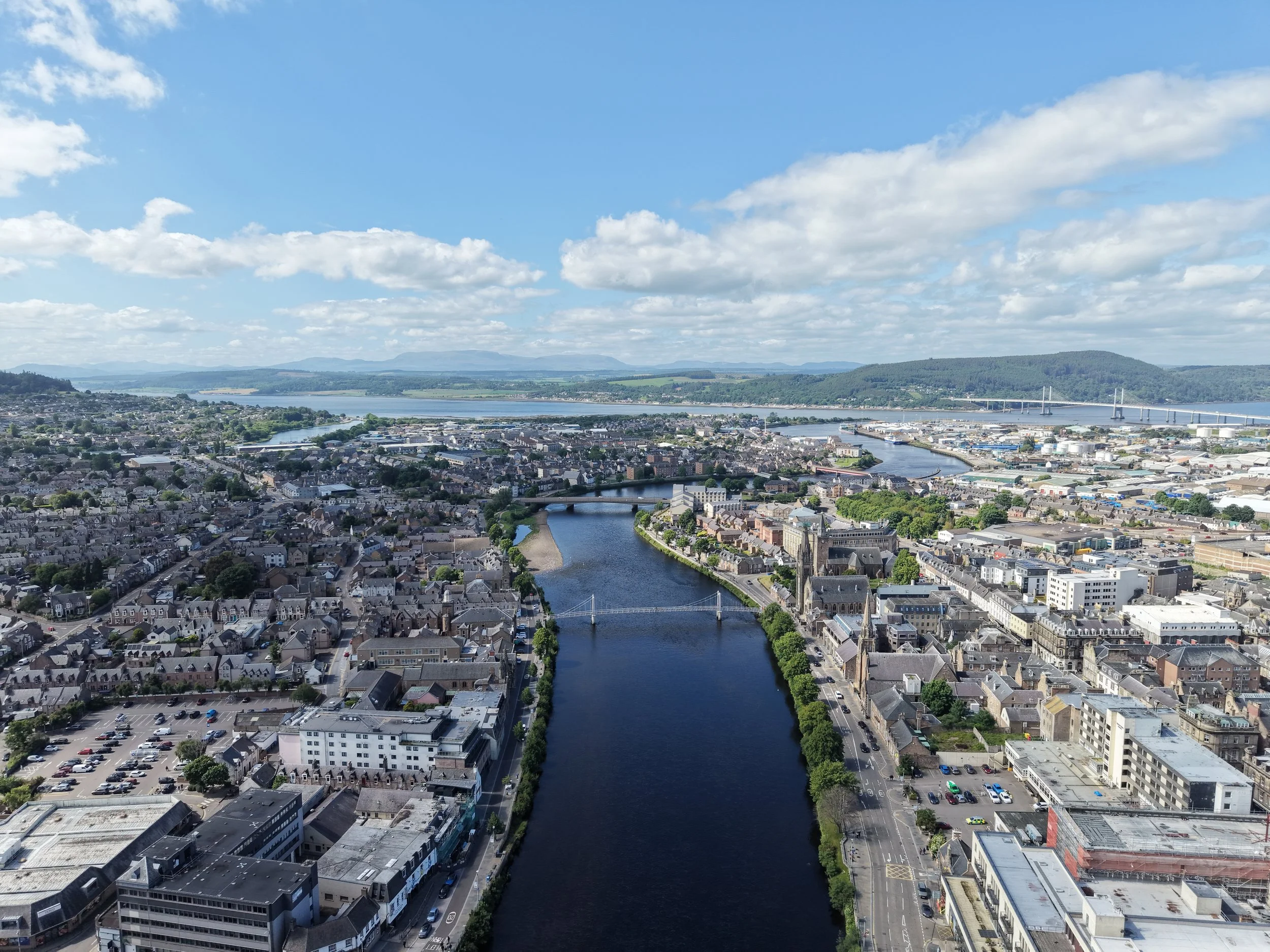 Aerial view of a city with a river running through it, surrounded by hills and a blue sky with scattered clouds.