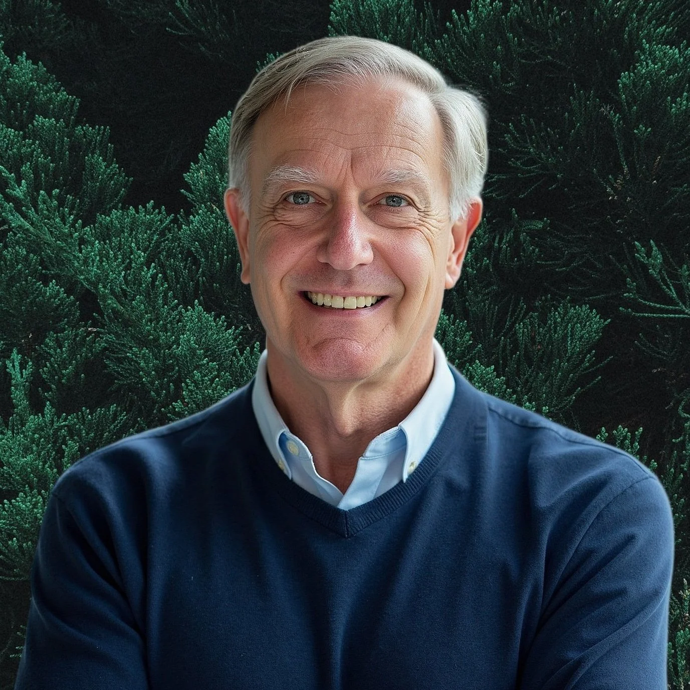 An older man with gray hair, smiling, wearing a navy blue sweater over a collared shirt, standing in front of a dense green pine tree background.
