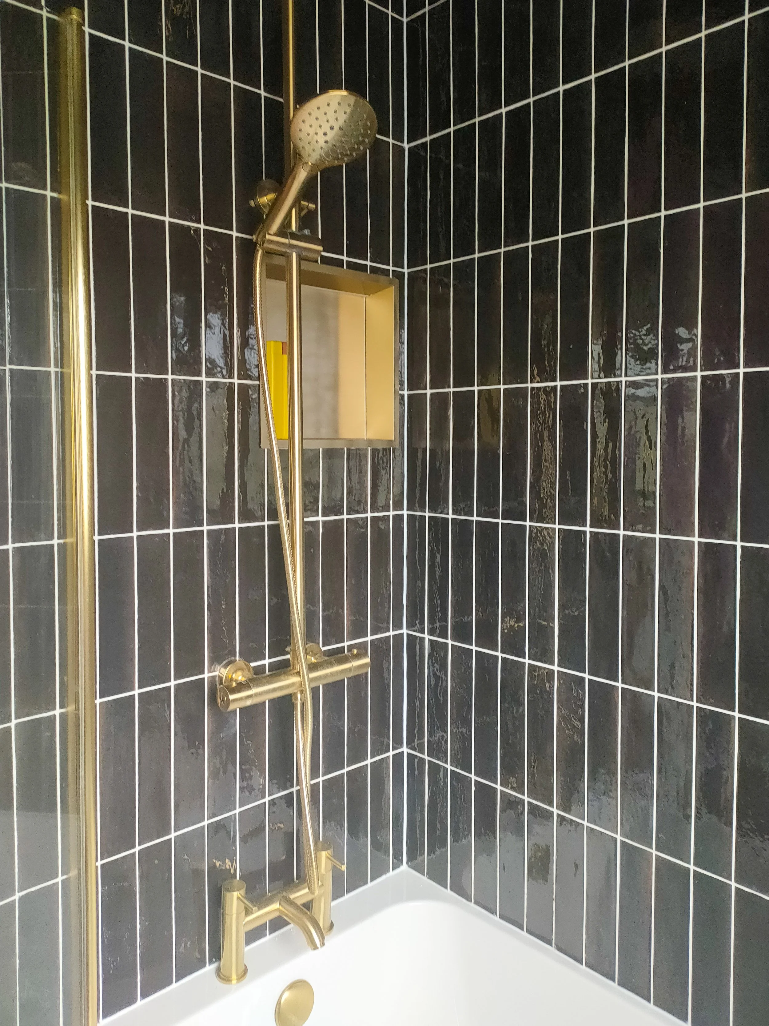 A modern shower with black tiled walls, a gold showerhead, and gold fixtures, with a glass shower door and a soap dish.