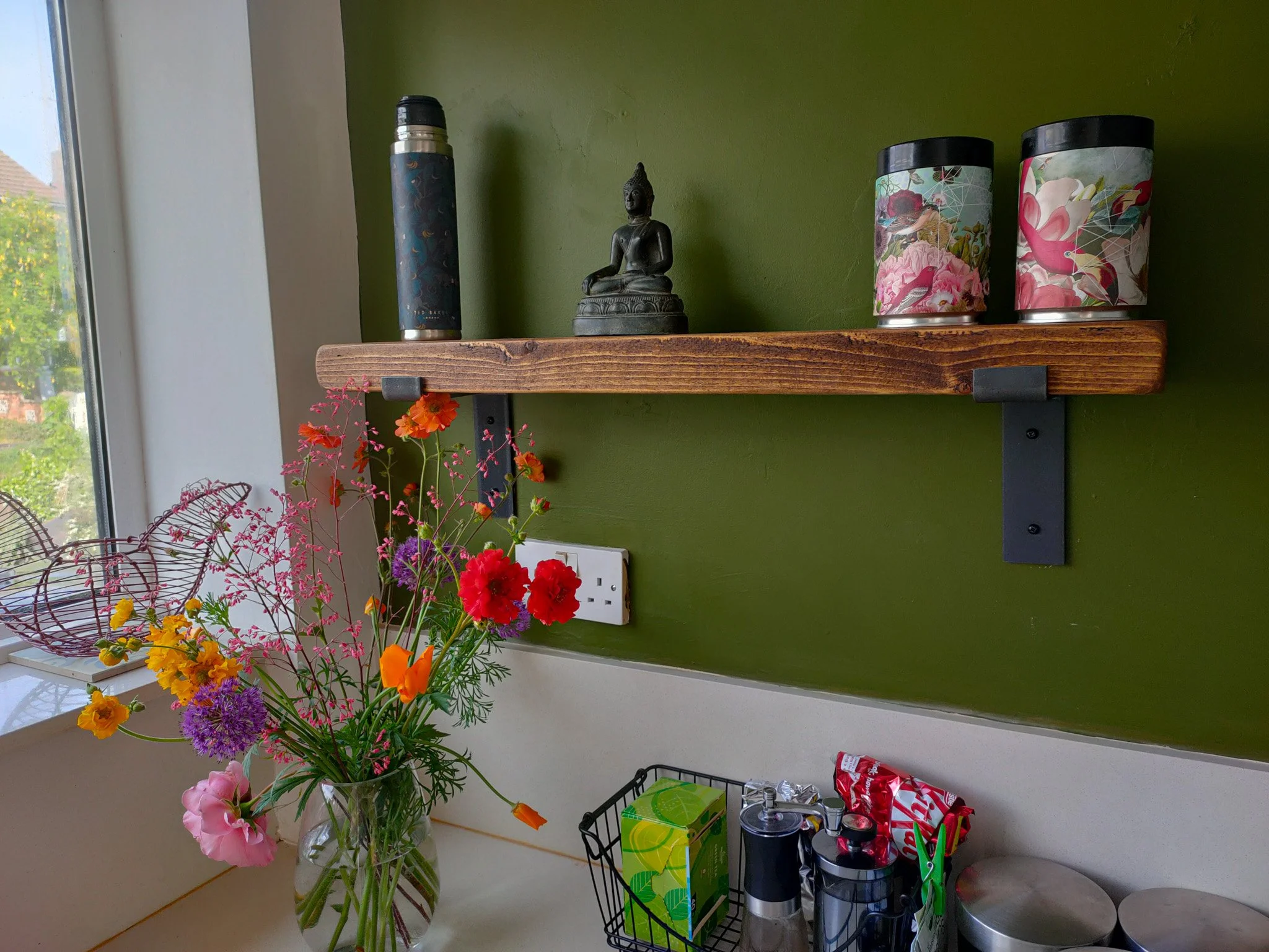 A kitchen windowsill decorated with a large glass vase of colorful flowers including pink, purple, orange, and red blooms, and a small pink flower arrangement. There are two large decorative tins with floral and flamingo designs on a green wall shelf, a vertical aluminum thermos, a black wire basket containing various packaged items, and a countertop with miscellaneous containers and snacks. The window shows a tree and some houses outside.