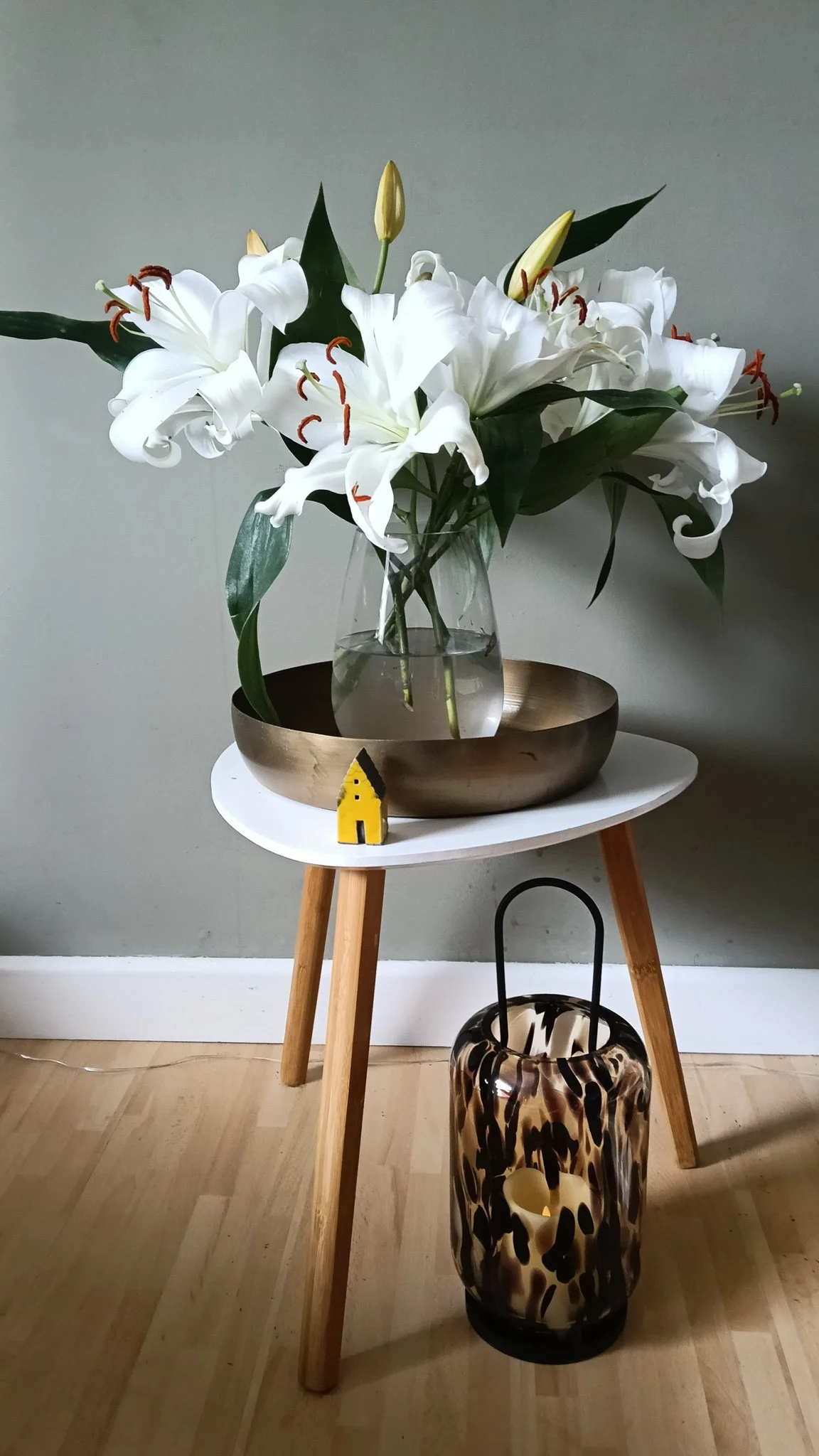 A vase of white lilies with green leaves on a small white table, with a yellow house-shaped object, a glass bowl, and a patterned lamp beside it on the floor, against a gray wall and wooden floor.