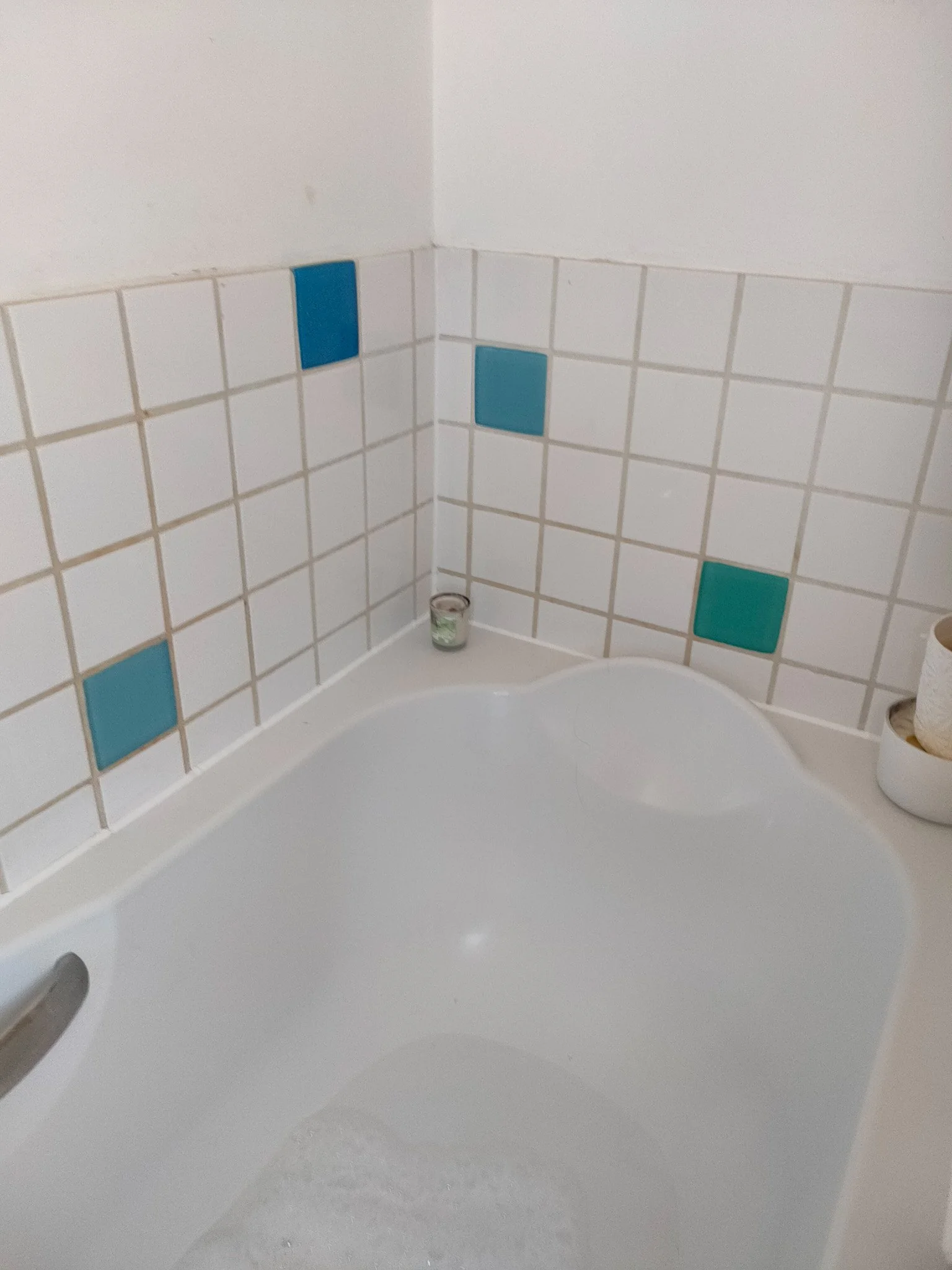 Close-up of a white bathtub with a tiled wall behind it, featuring a few blue accent tiles, a small candle on the edge, and a toilet paper roll in the corner.