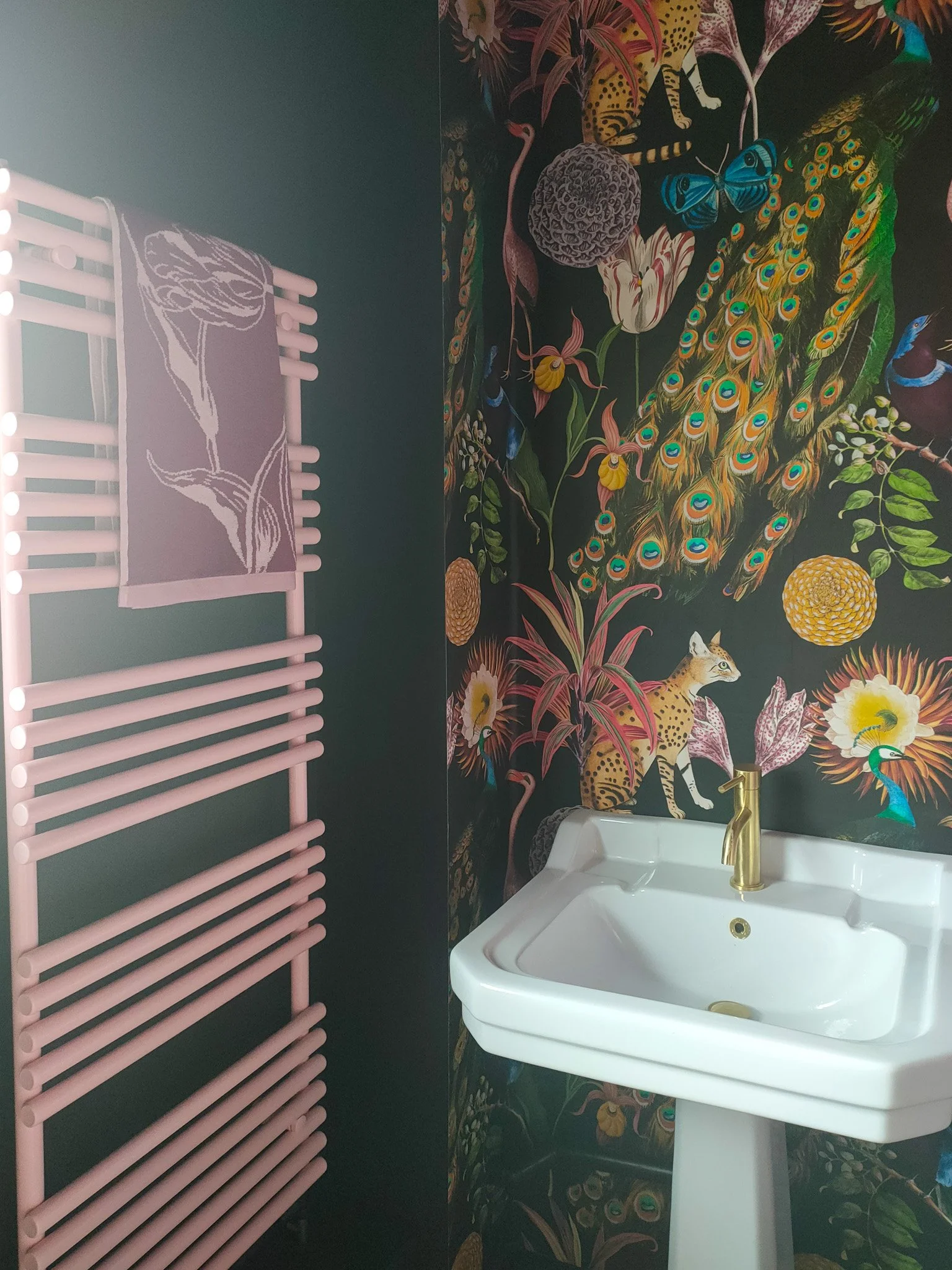 A bathroom wall decorated with vibrant floral and animal patterns, featuring a white sink with a gold faucet, a pink towel rack, and dark-colored walls.