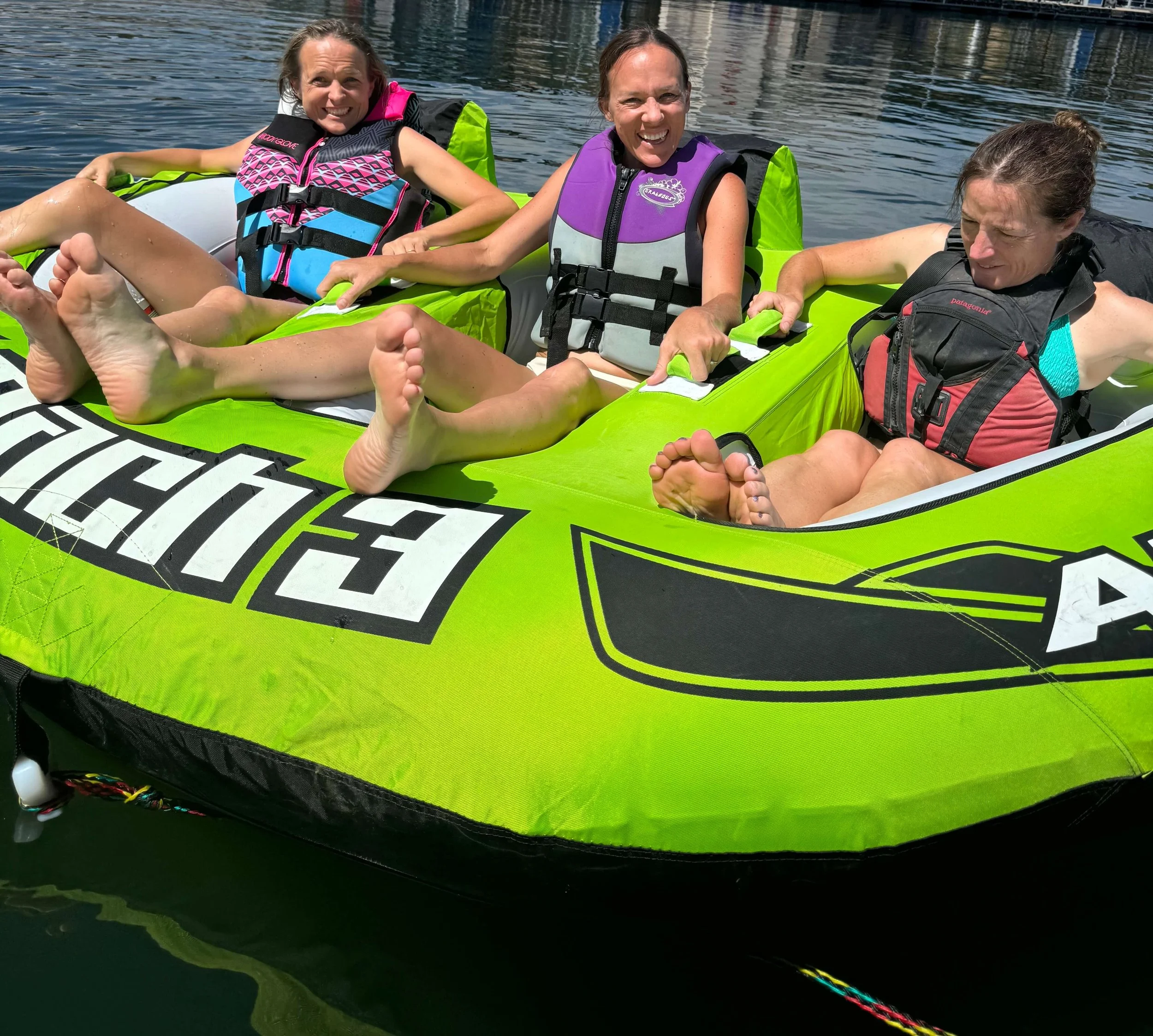Three women are sitting on a lime green inflatable raft on a body of water, all wearing life jackets and smiling at the camera.