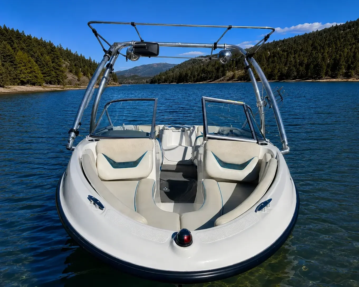 A white speedboat with a wakeboard tower, floating on a calm lake surrounded by forested mountains under a clear blue sky.