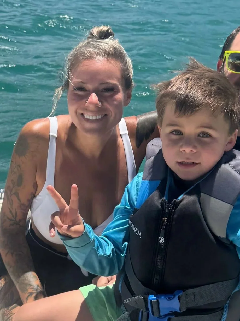 Smiling woman with tattoos on her arms, wearing a white swimsuit, on a boat with a young boy in a life jacket, making a peace sign, all on the water.