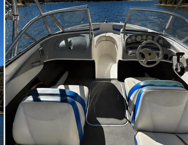 Interior of a boat cockpit with two white captain chairs, steering wheel, and dashboard, on a lake.