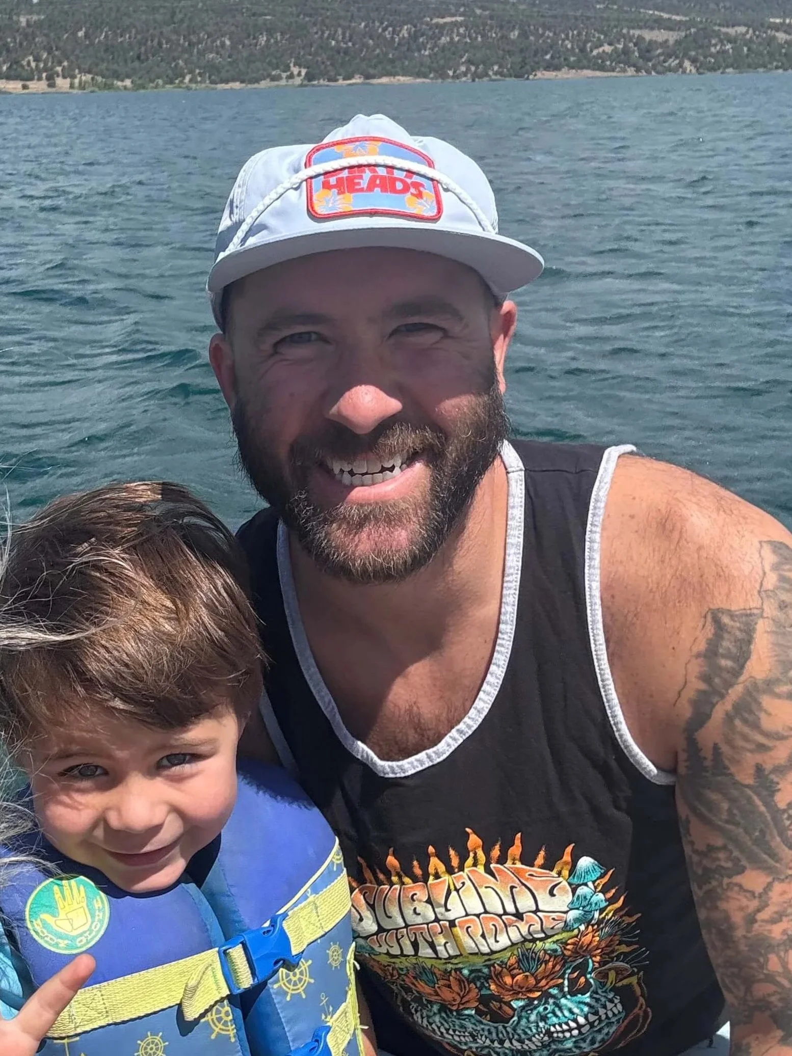 A man with a beard wearing a white cap with colorful text, smiling, outdoors on a body of water with a wooded shoreline in the background. A child with brown hair and a blue life jacket is partially visible in the foreground, pointing with a finger.