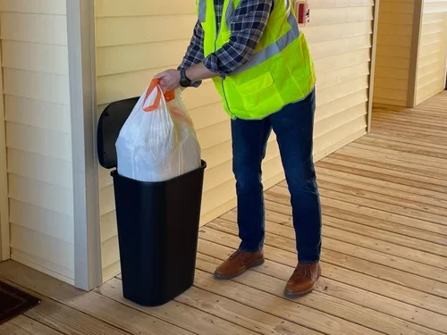 Person disposing of trash into a black trash can on a wooden porch, wearing a yellow safety vest and blue jeans.