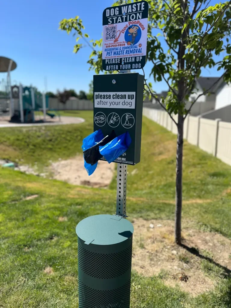 Dog waste station with a bin and signage, including a bag dispenser, on a grassy area with trees and houses in the background.