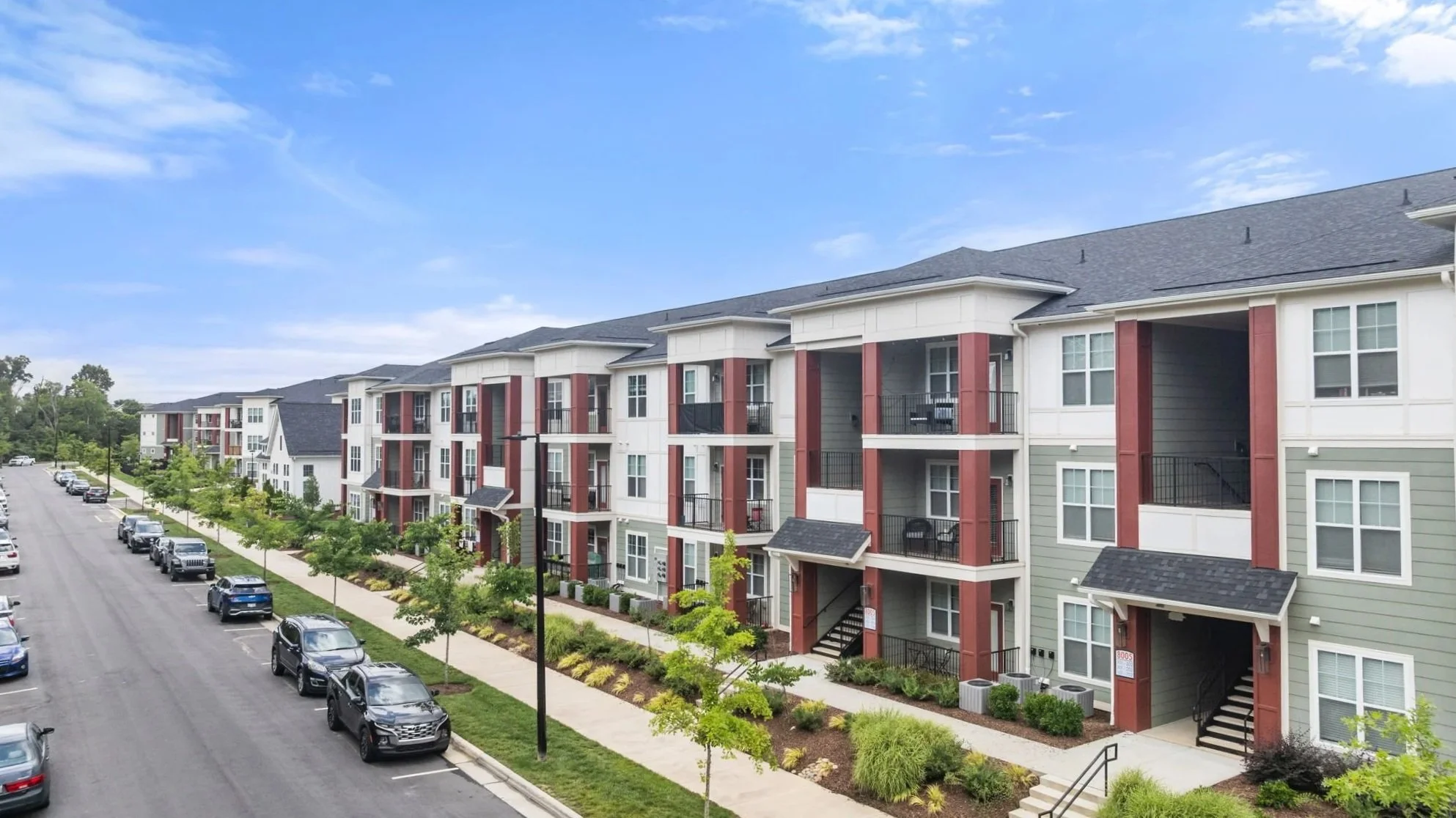 A multi-story residential apartment complex with balconies, green landscaping, and parking lot on a sunny day.