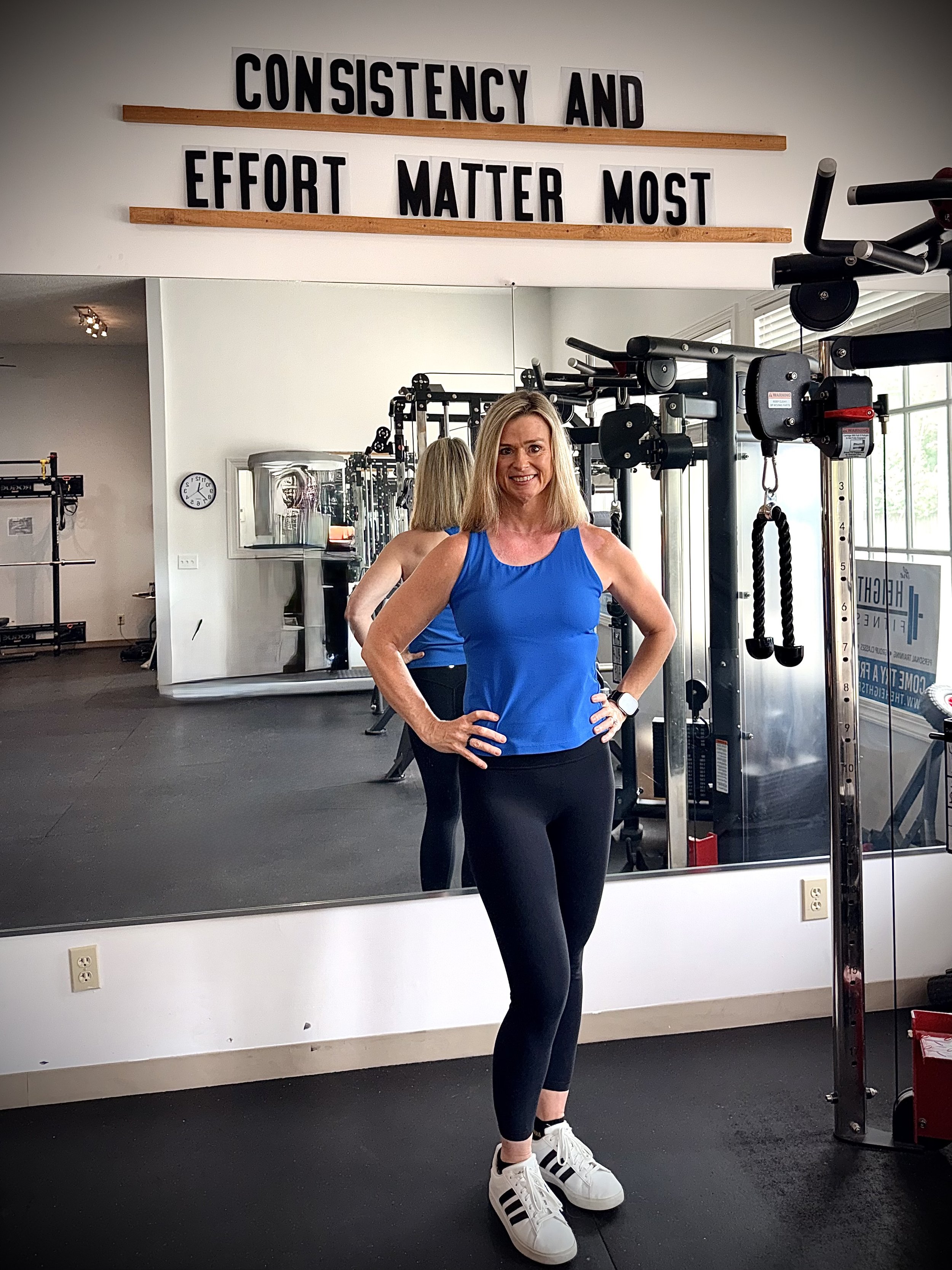 A woman in a blue tank top and black leggings posing in front of a gym mirror, with gym equipment behind her and motivational signs above.