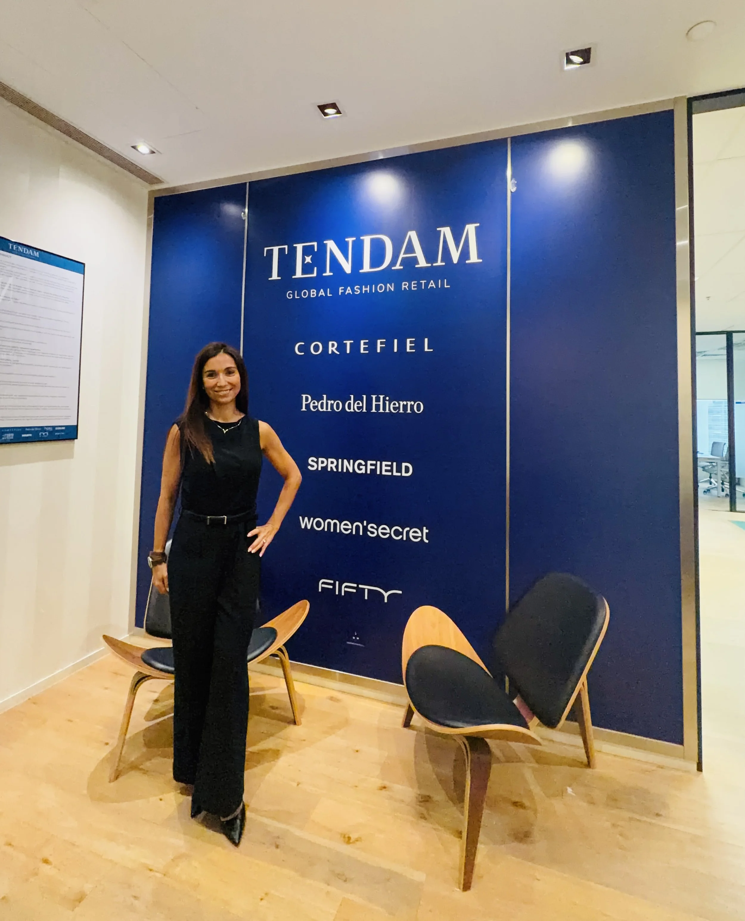 Woman in black jumpsuit in front of a company backdrop in blue with brand names in white from TENDAM group