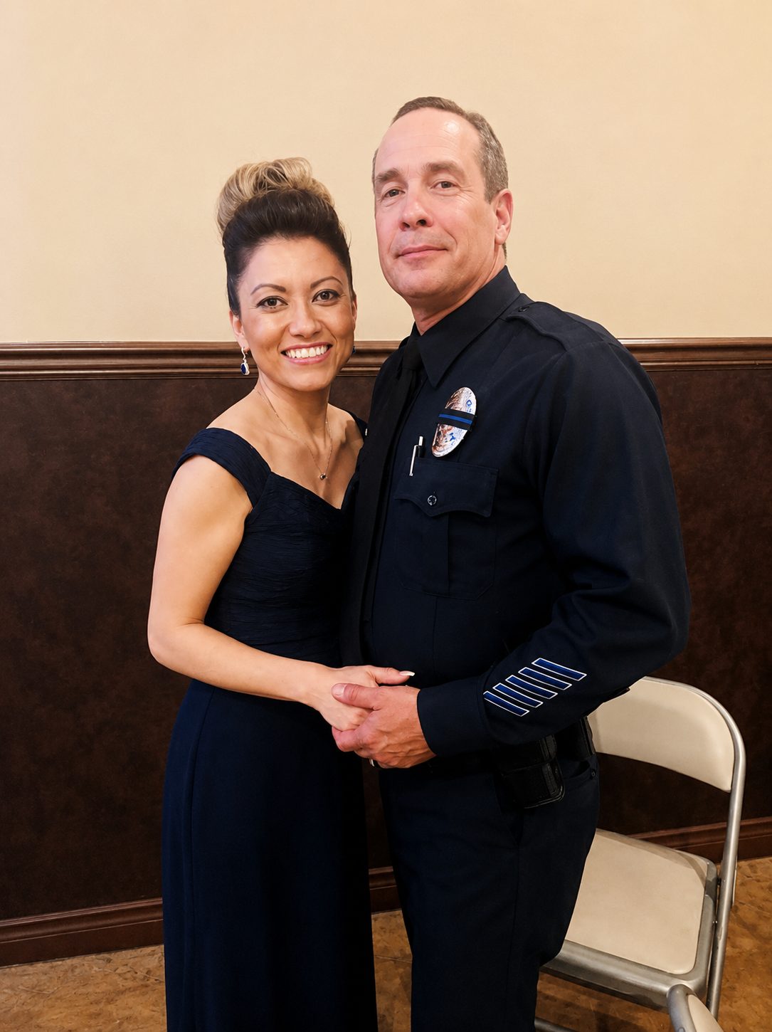 Rich and Amanda Rodriguez holding hands and posing together, both smiling in front of a plain beige and brown wall.