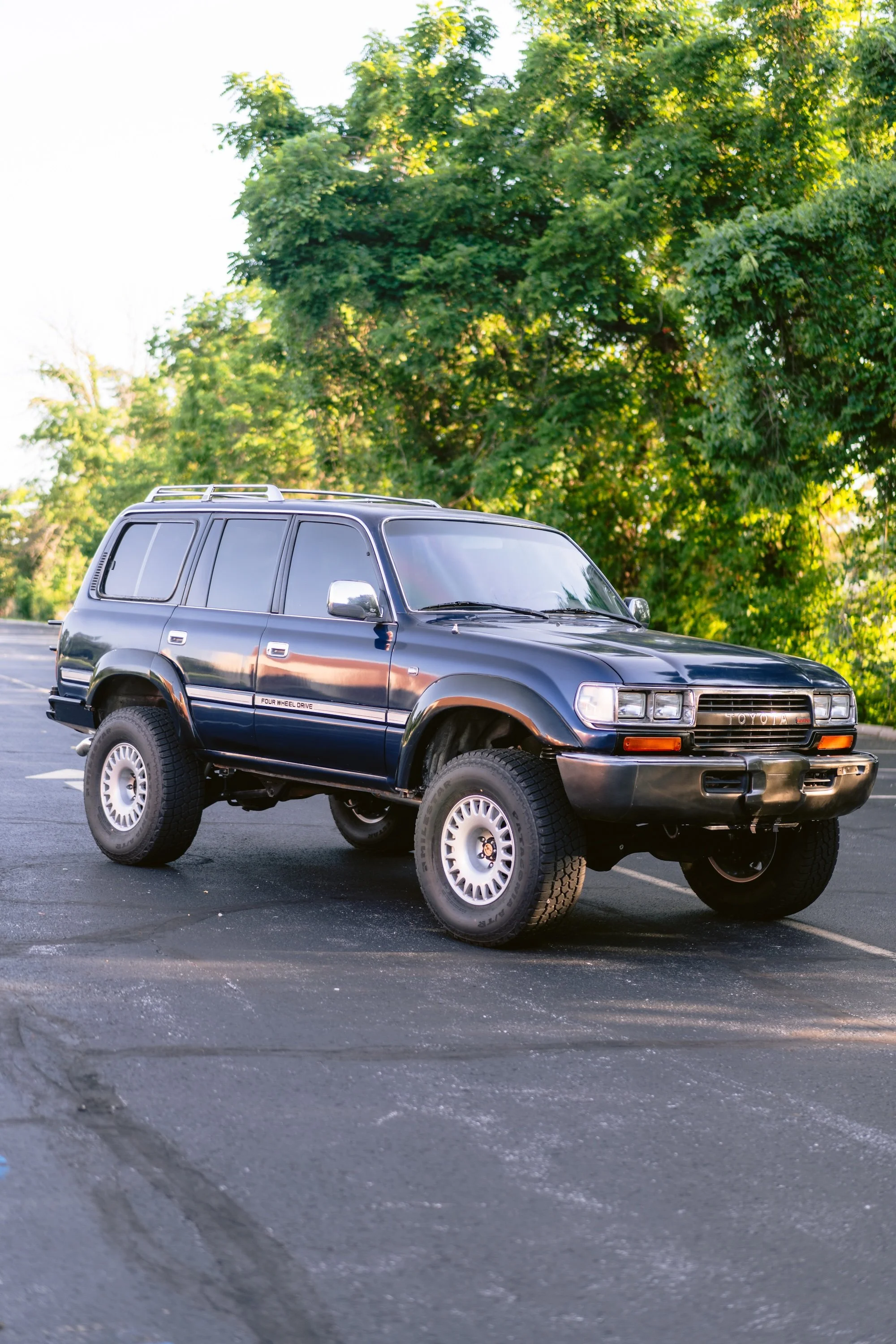 A black Toyota SUV with large off-road tires is parked in an outdoor parking lot with a background of green trees and sunlight.